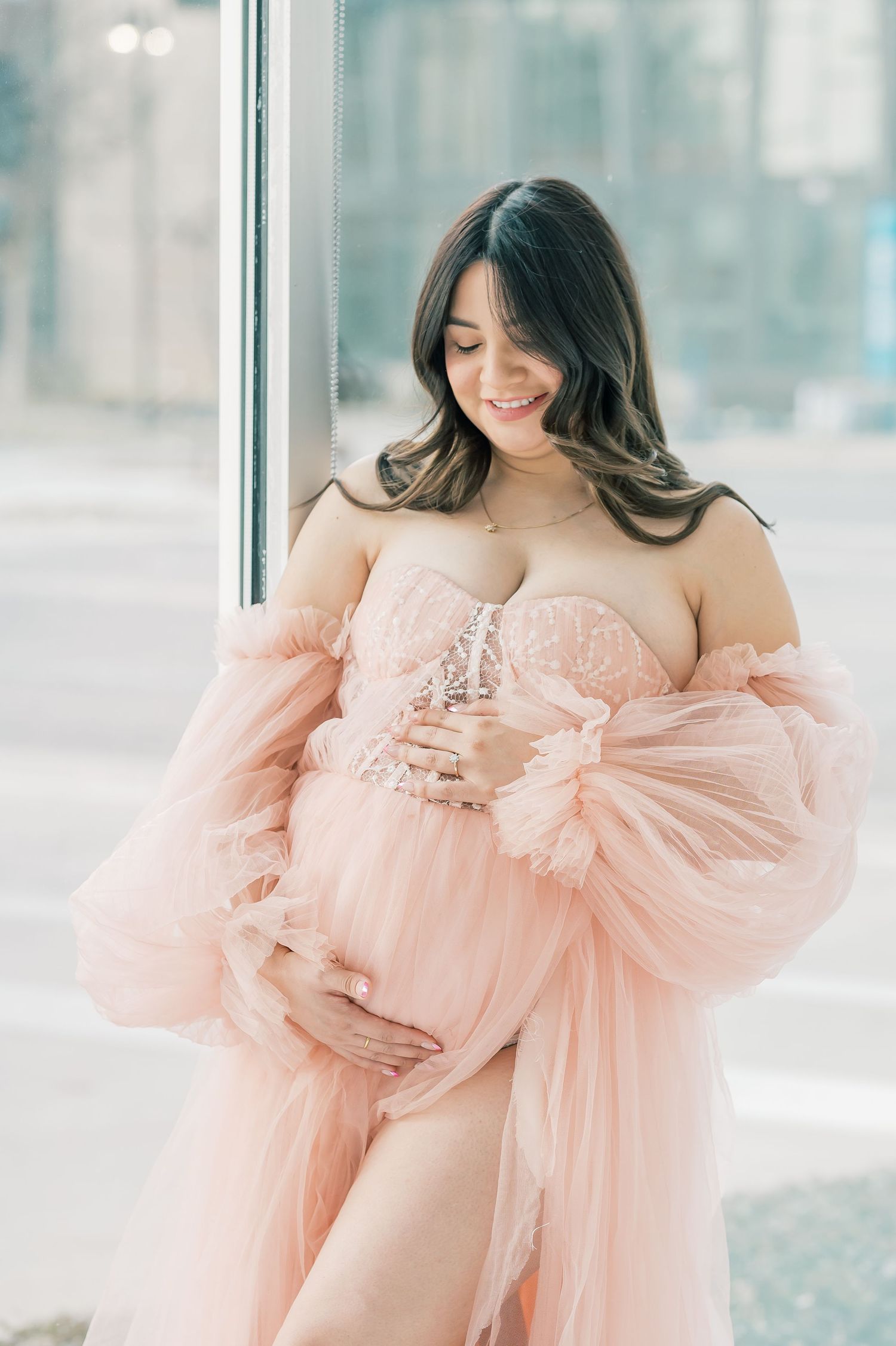 Pregnant woman in pink tulle robe and lace bodice cradling baby bump near window.