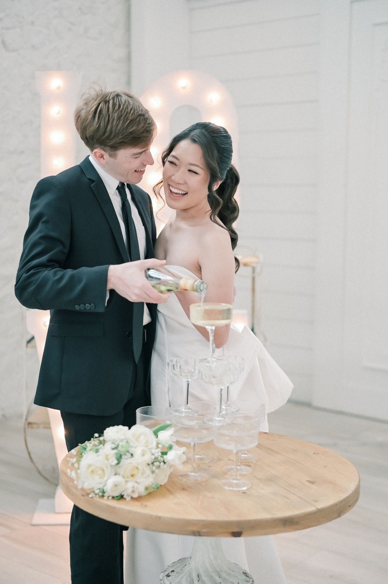 Bride and groom smiling while pouring champagne tower at wedding reception with glowing LOVE sign backdrop.