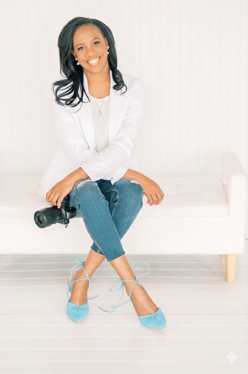 Smiling woman in white blazer and jeans sits cross-legged holding a camera, wearing teal heels on a white couch.