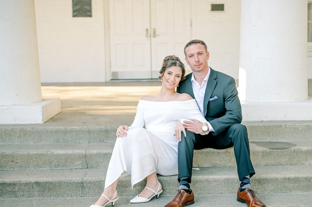 Stylish couple in white and navy sit together on stone steps of a classic white building for maternity photos.