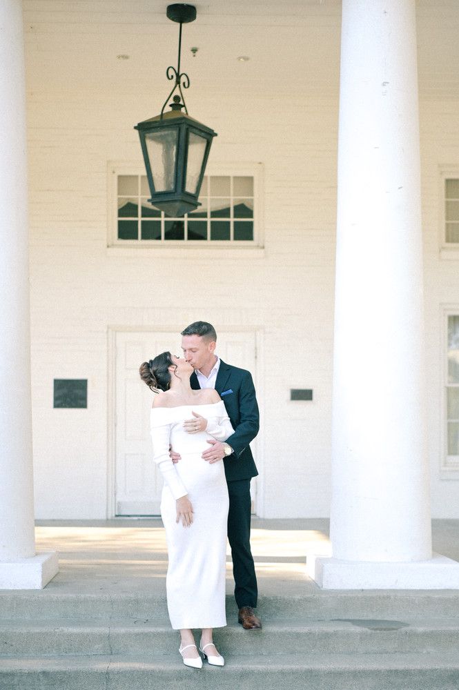 Couple sharing a romantic kiss on white columned porch steps, bride in elegant off-shoulder white gown, groom in navy suit.
