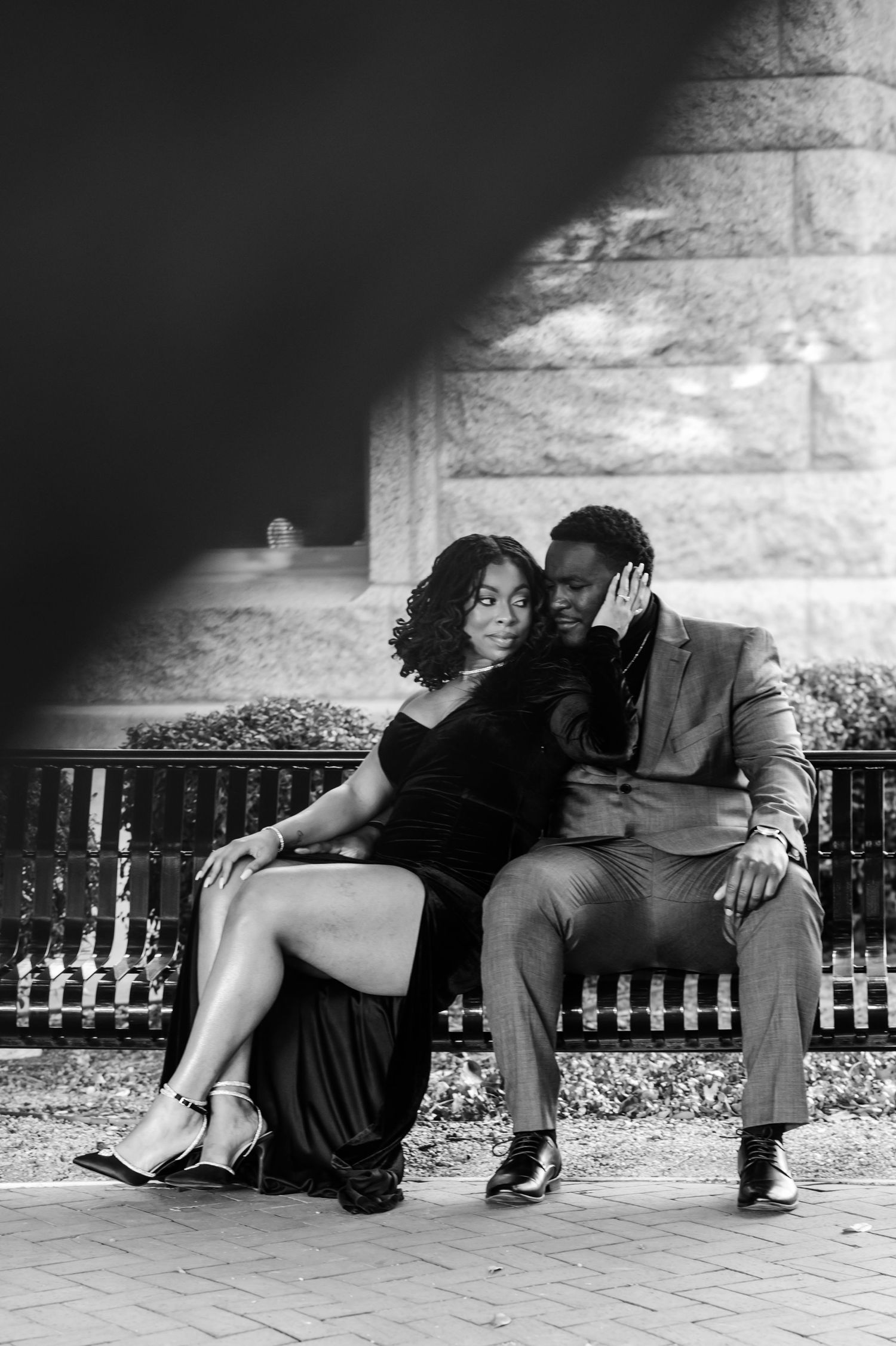 Black and white photo of a couple sitting intimately on a bench, woman in black dress leaning on mans shoulder outdoors.