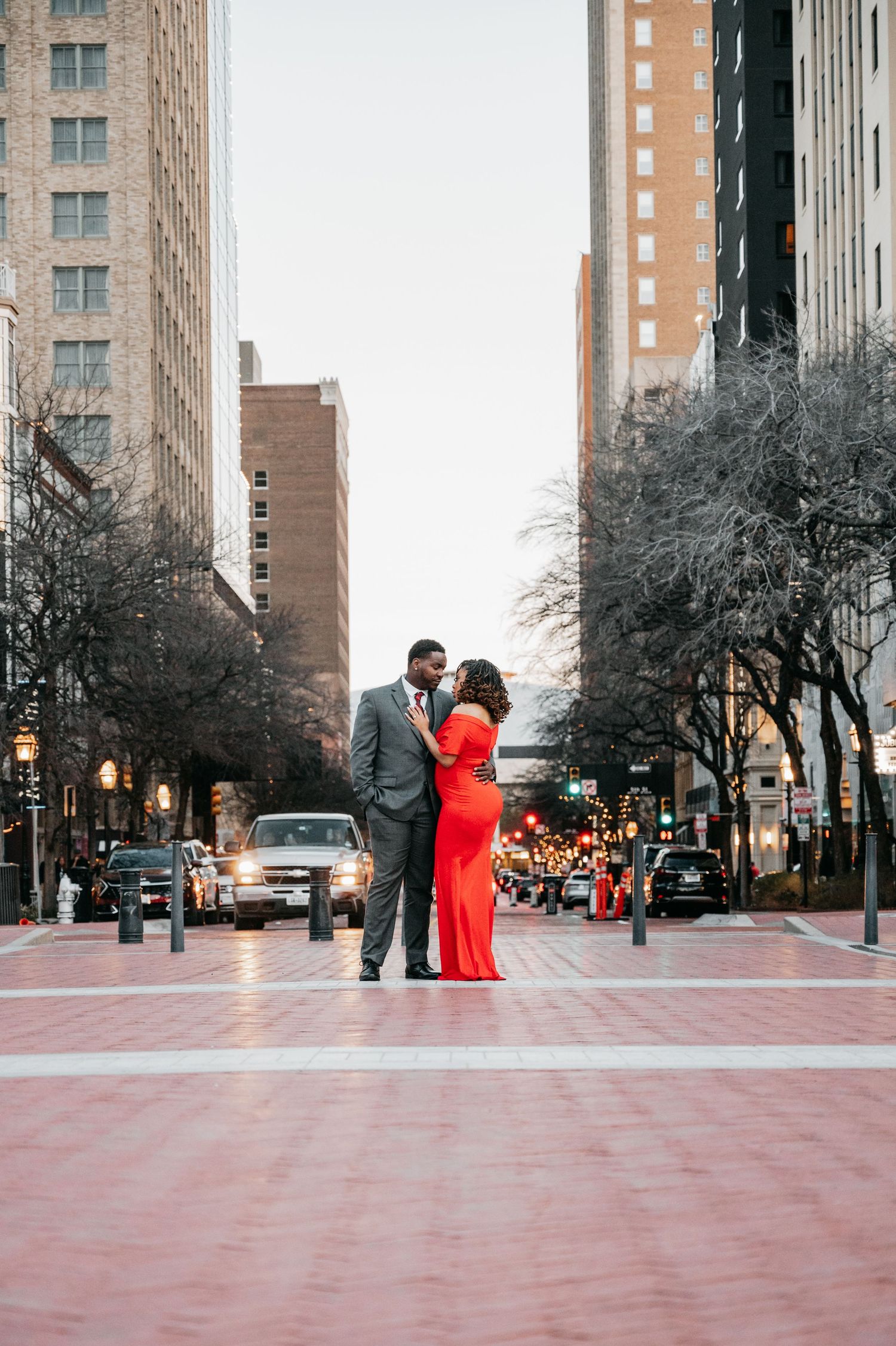 A couple shares a romantic moment on a city street, woman in red gown, man in gray suit, urban skyline backdrop.