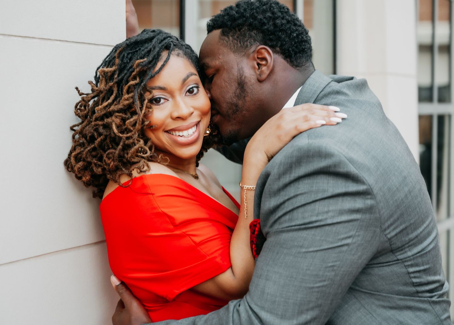 A man in a gray suit kisses a smiling woman in a red dress on the cheek during a romantic engagement photo.