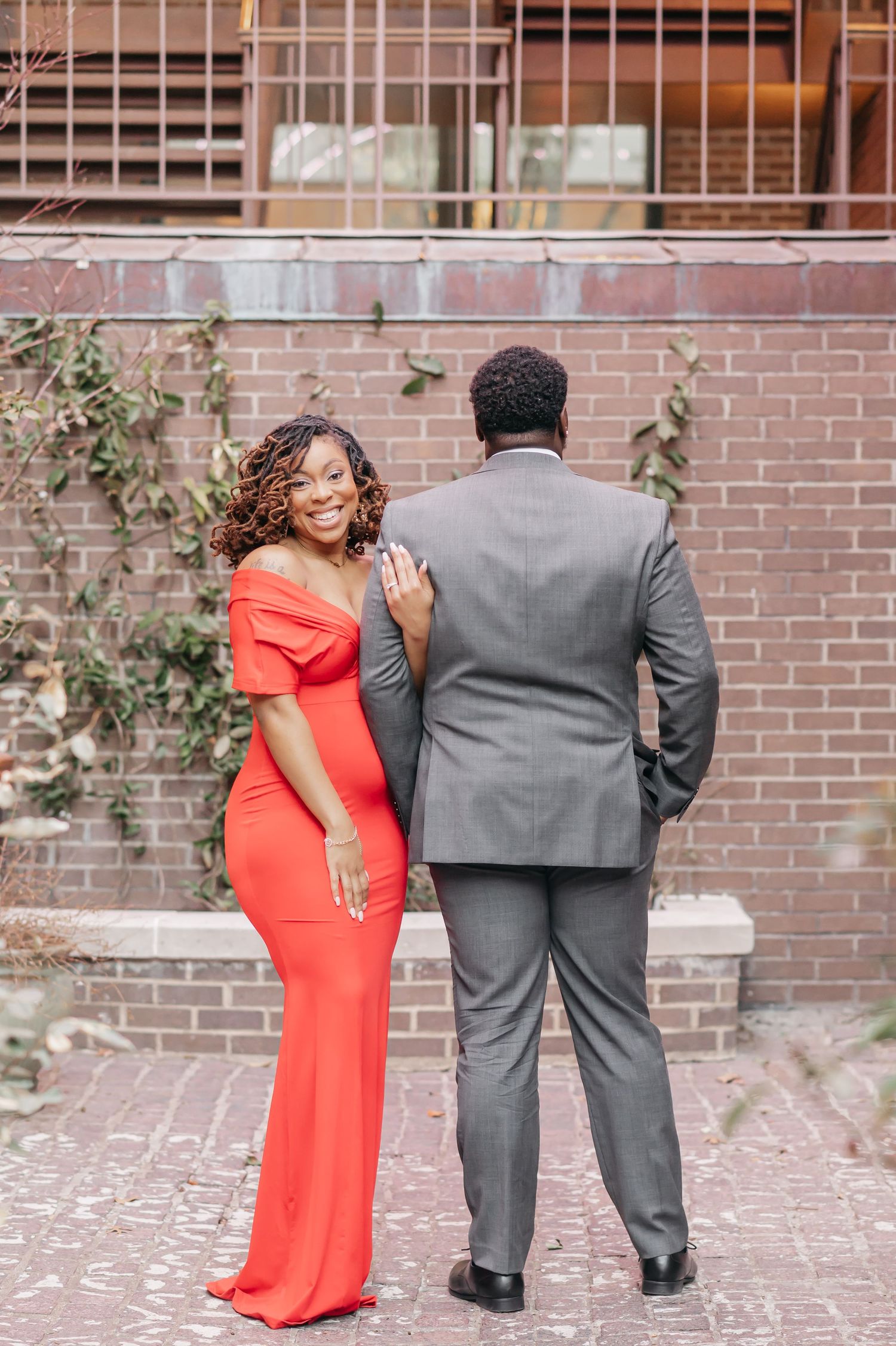 Smiling woman in red off-shoulder gown poses with man in gray suit against brick wall during engagement shoot.