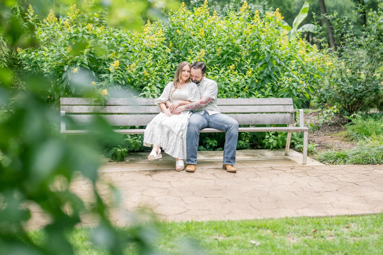 A couple sits closely together on a wooden bench surrounded by lush green garden foliage.