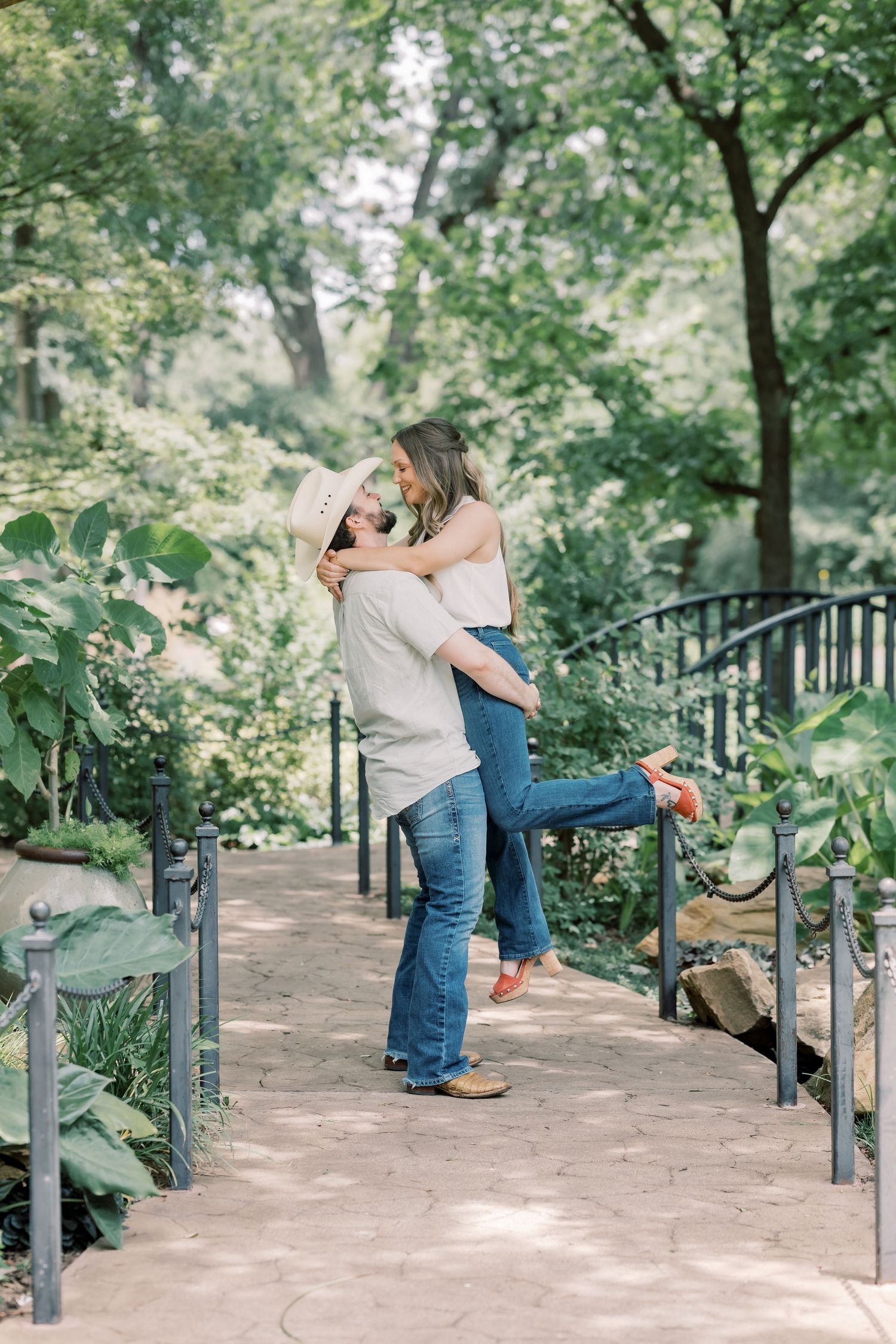 Man in cowboy hat lifting woman in jeans and white top on a garden path surrounded by lush green trees.