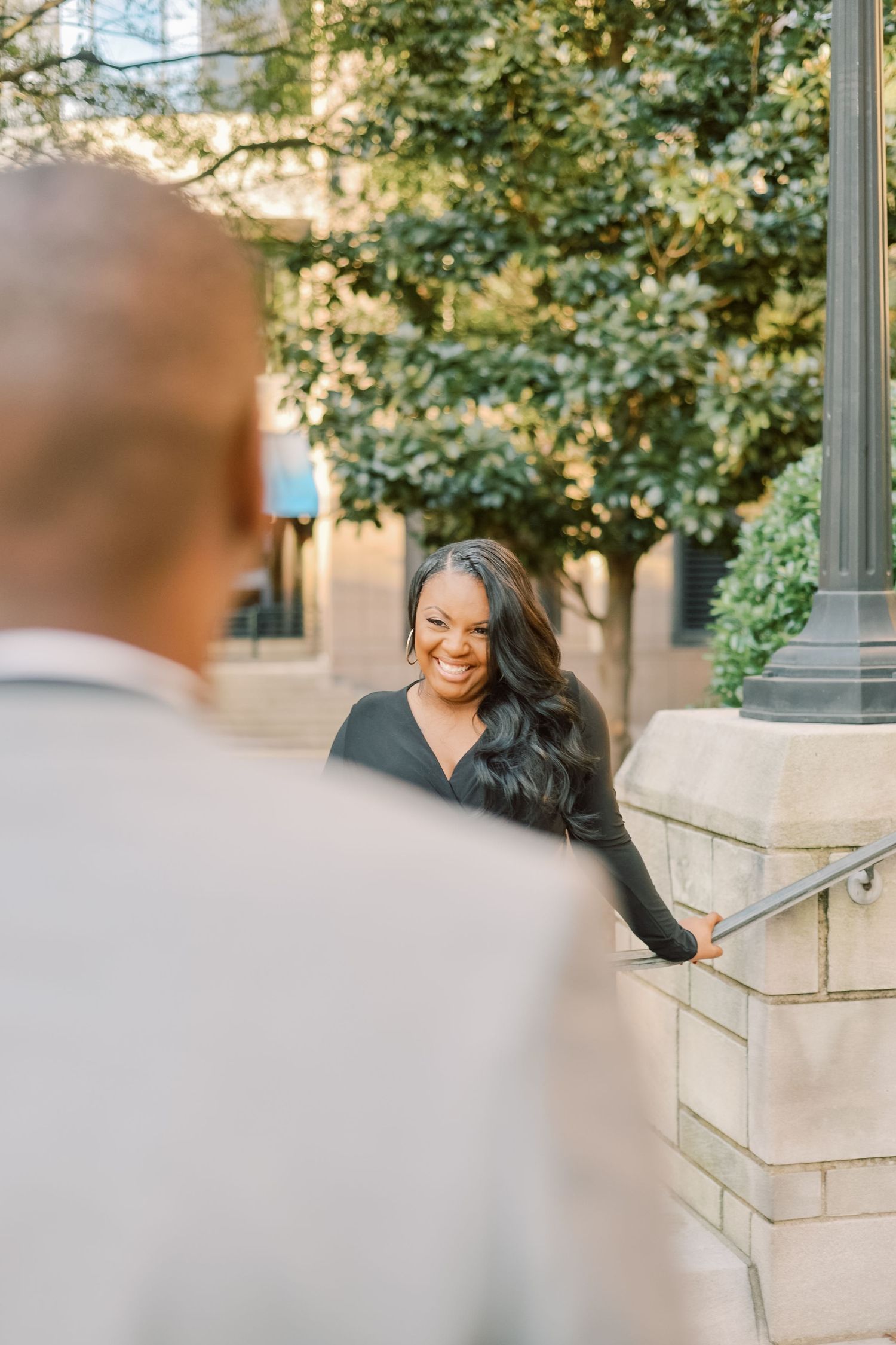 A smiling Black woman in a black top leans on a railing outdoors, looking toward a man in a light suit.