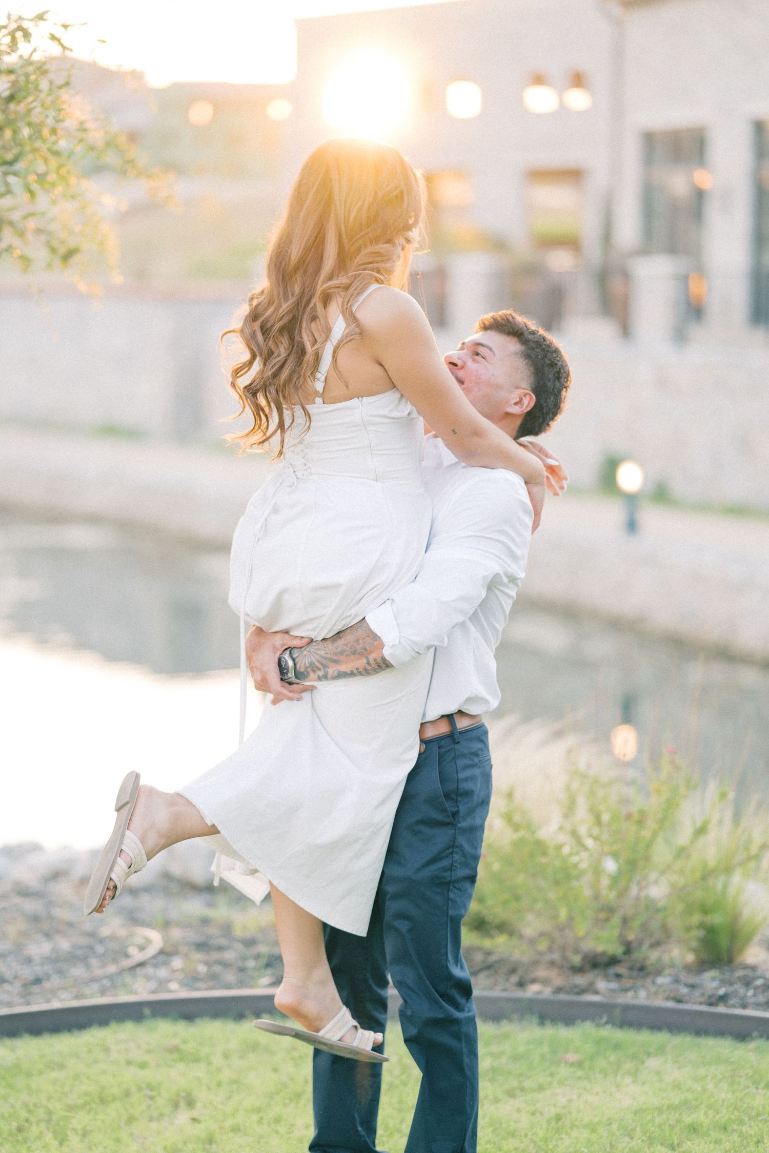 Man lifting woman in white dress during glowing golden sunset engagement photo session.