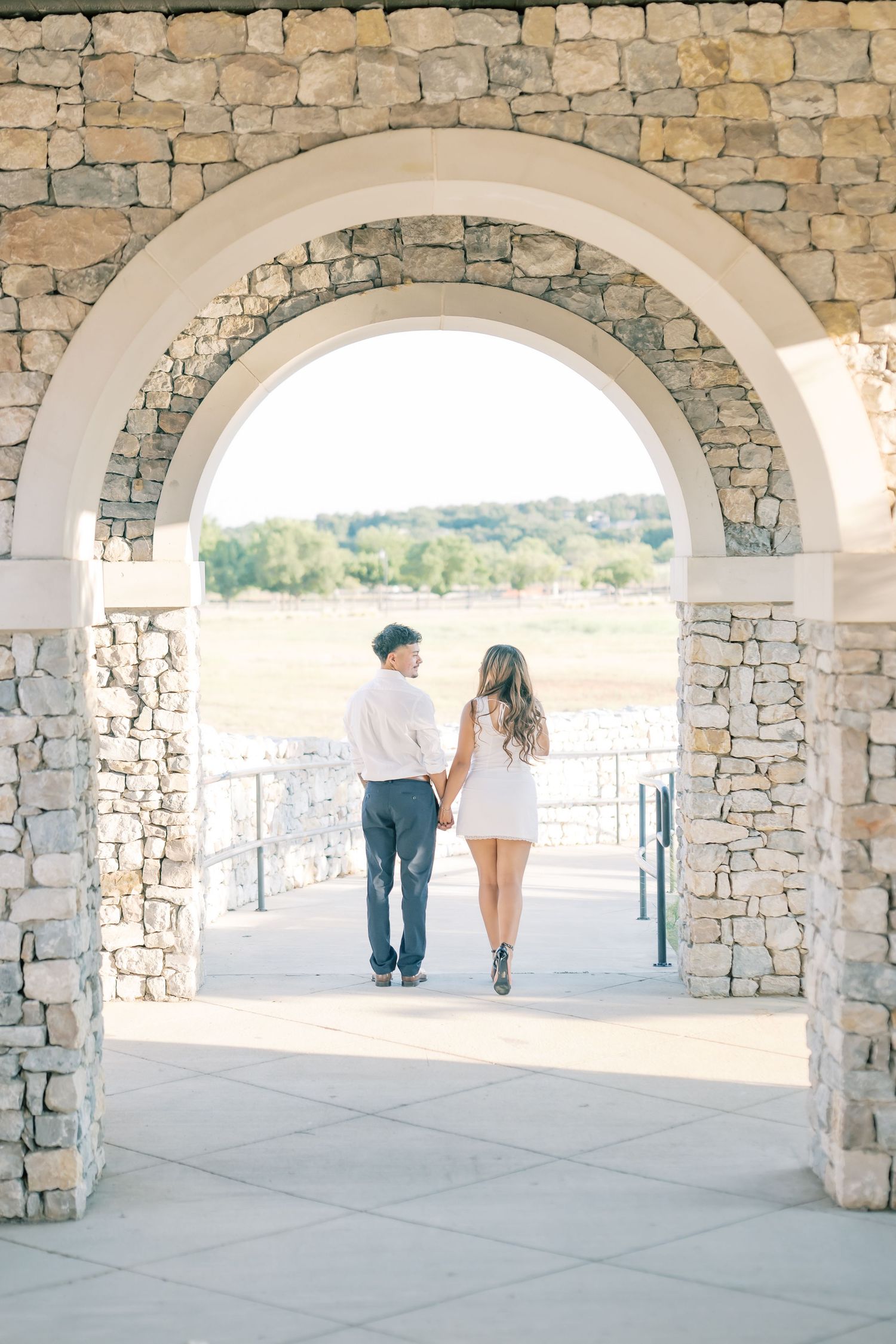Engaged couple walking hand in hand through a rustic stone archway at sunset.
