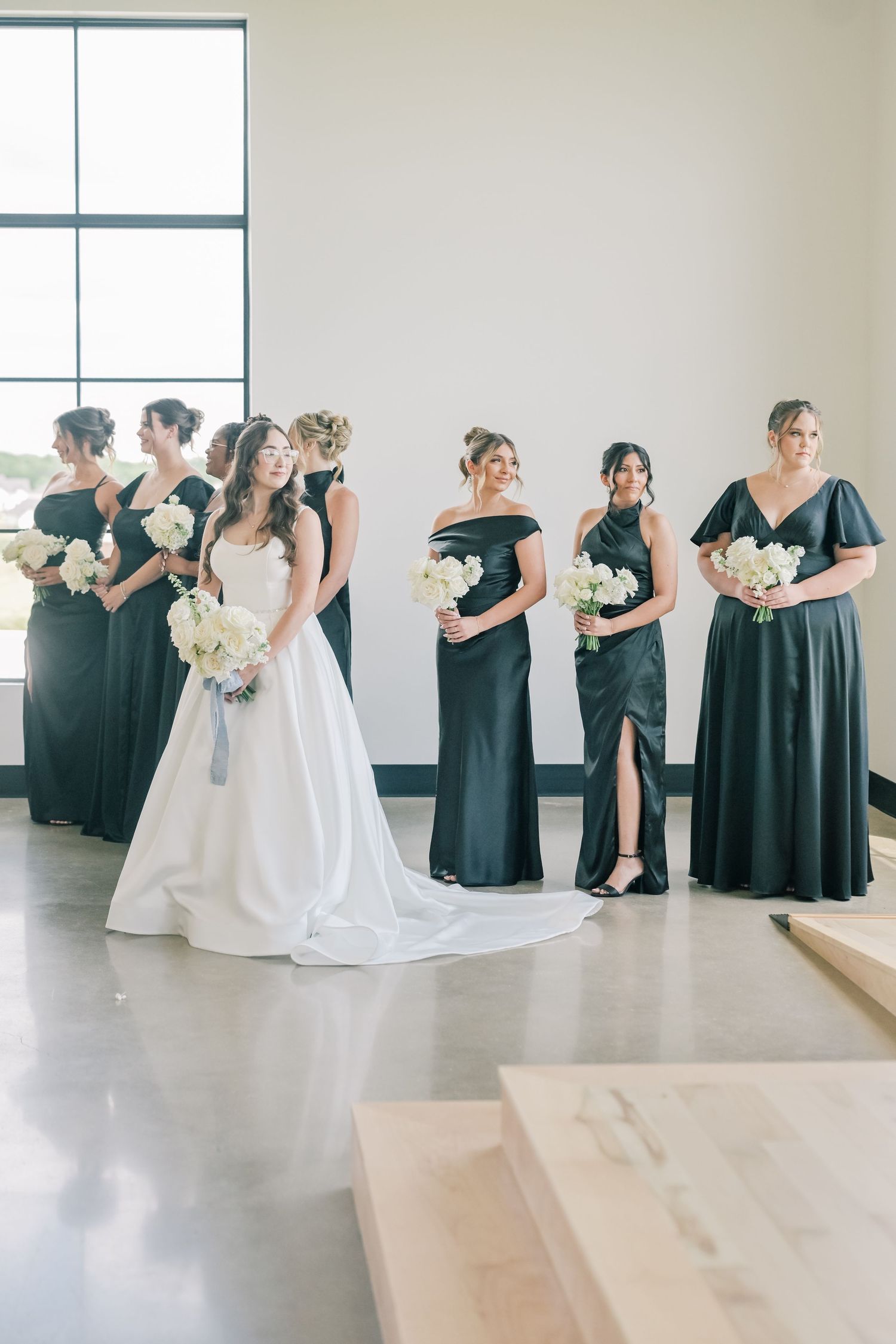 Bride in a white ball gown stands with bridesmaids in dark navy dresses holding white floral bouquets.