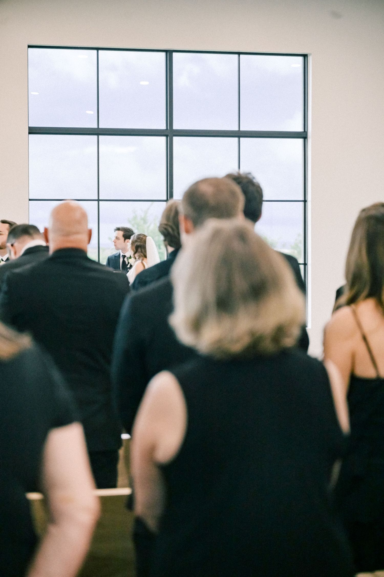Wedding guests in black attire stand watching the ceremony near a large window in a modern venue.