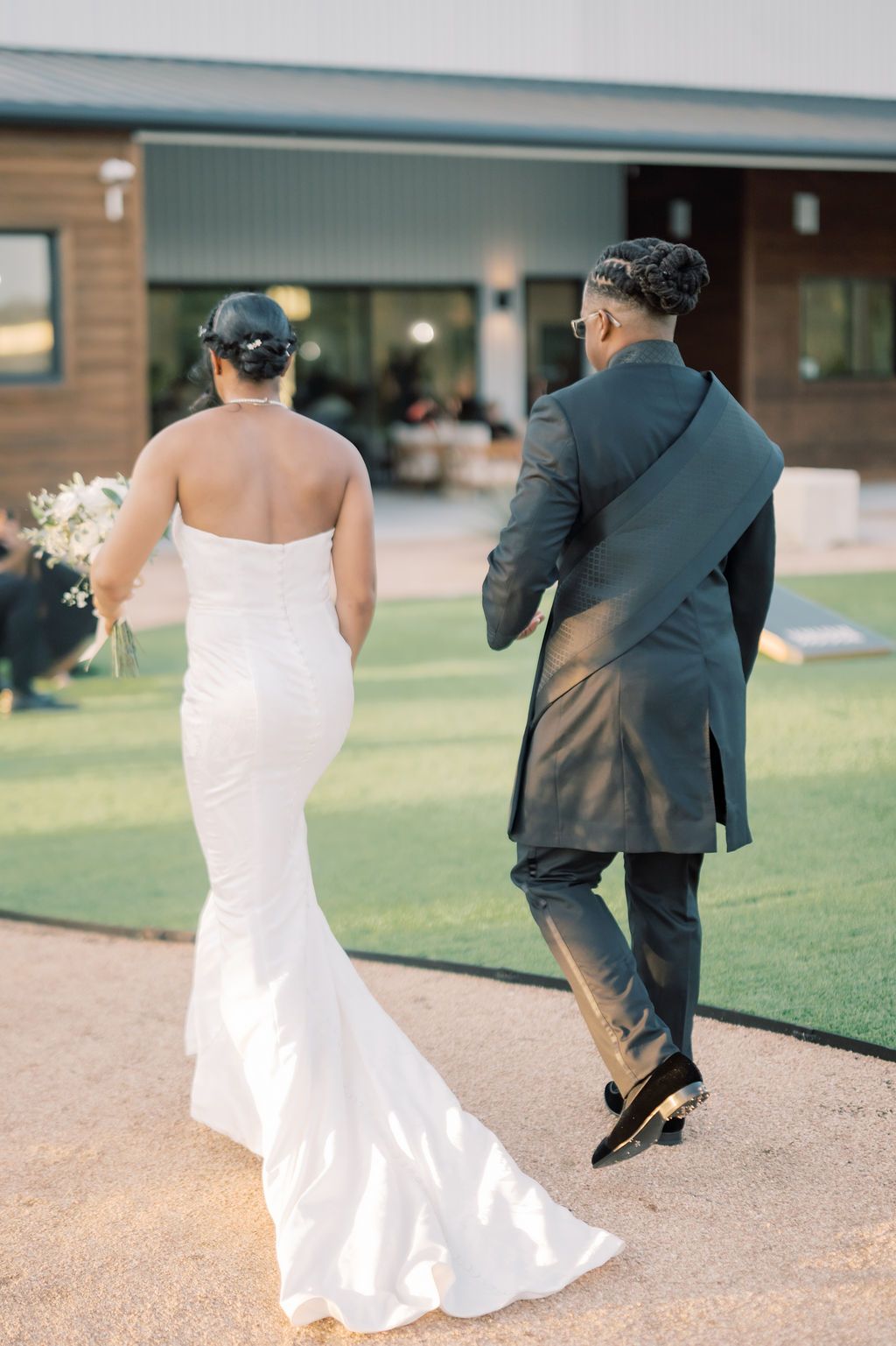 Bride in white mermaid gown and groom in dark suit walk hand-in-arm toward venue on their wedding day.