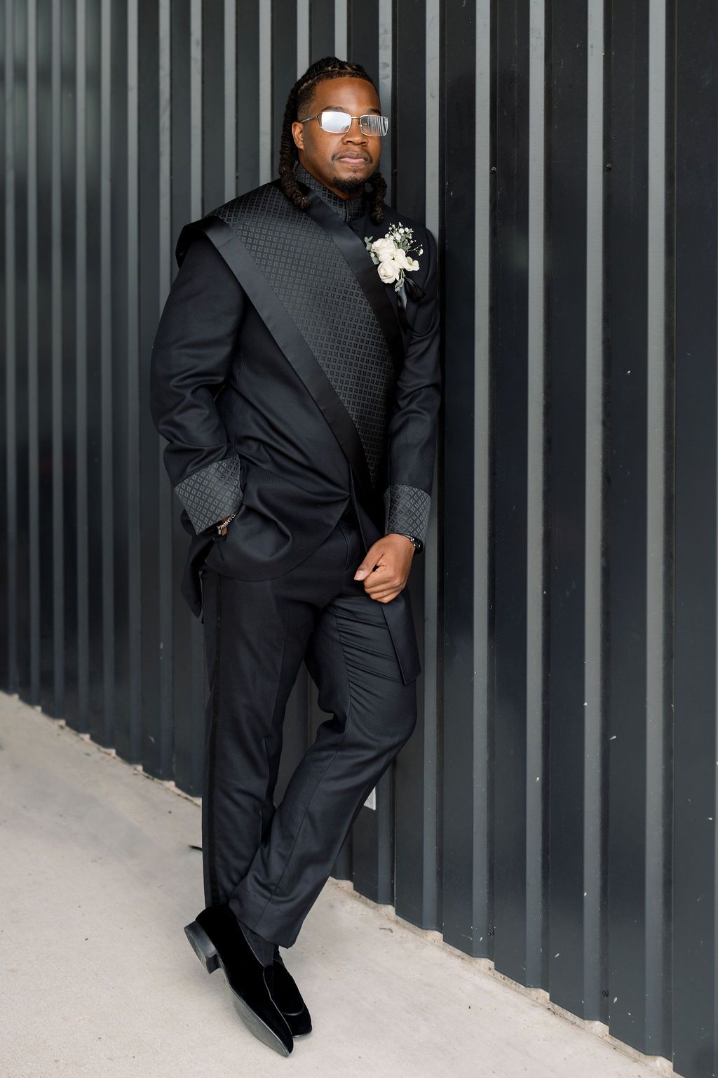 Stylish groom in unique black tuxedo with white boutonniere and silver sunglasses posing outdoors.