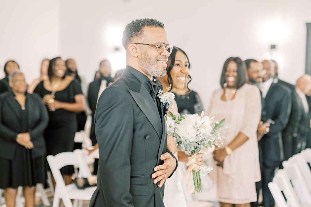 Bride walks down the aisle with escort during elegant indoor wedding ceremony with guests in black attire.
