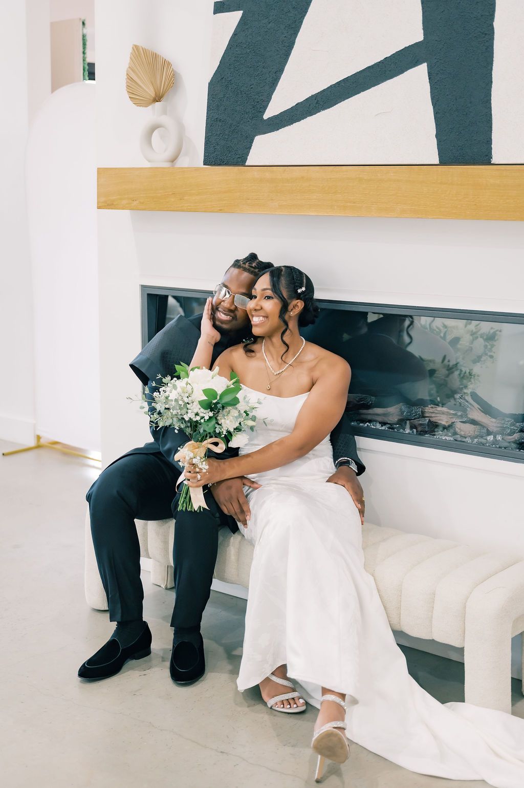 Bride and groom sit together by a modern fireplace, bride holding white bouquet in elegant white gown.