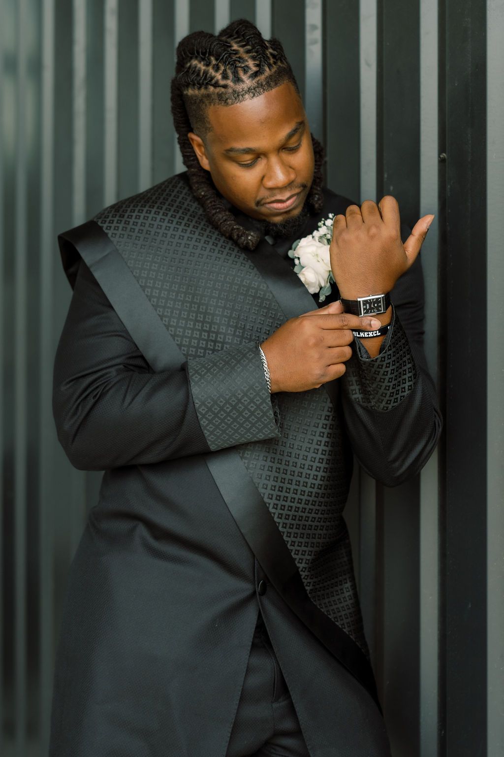 A well-dressed Black man in a black tuxedo with braided hair adjusts his watch, wearing a white boutonniere corsage.
