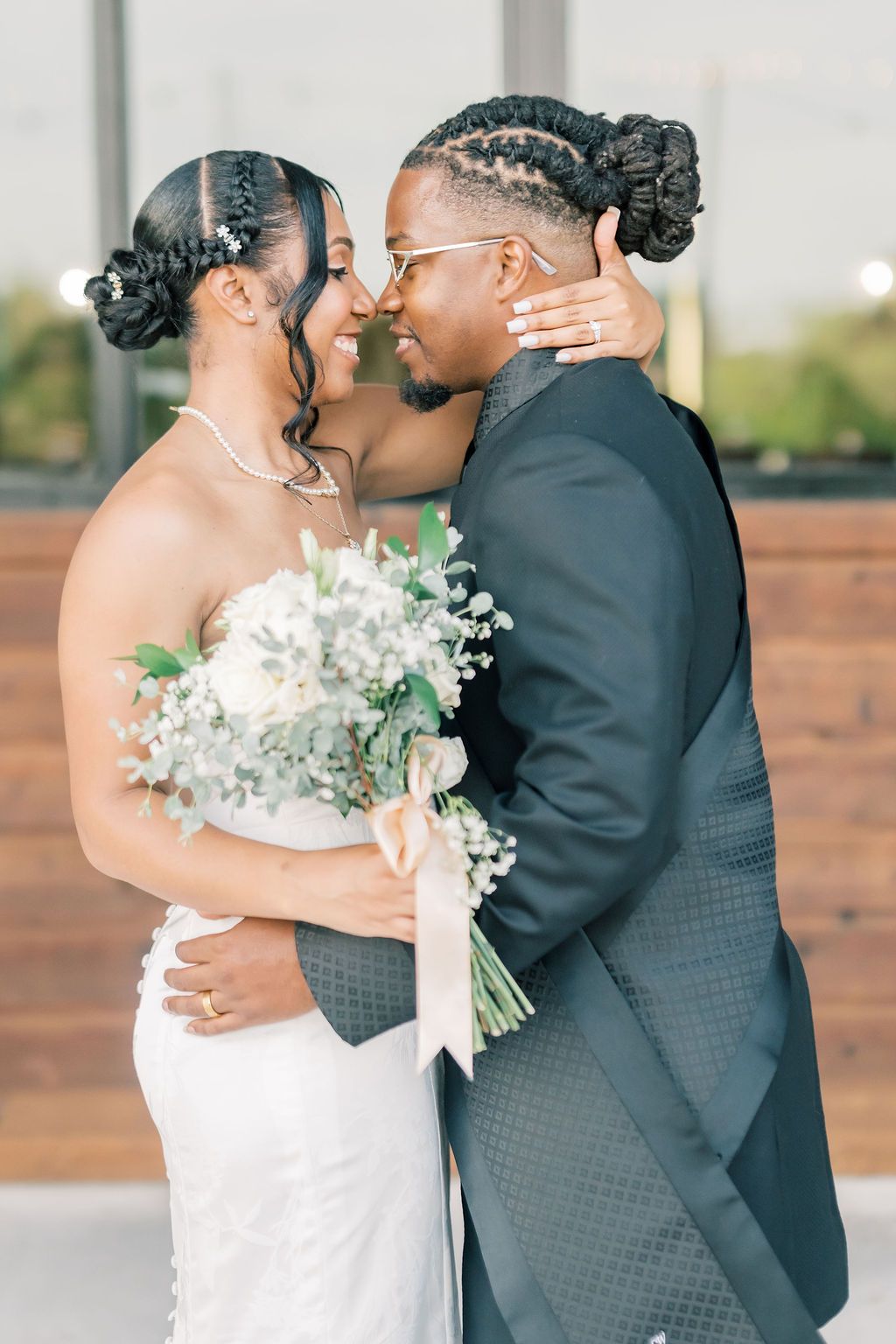 A joyful Black bride and groom in wedding attire share a romantic moment, holding a white floral bouquet.