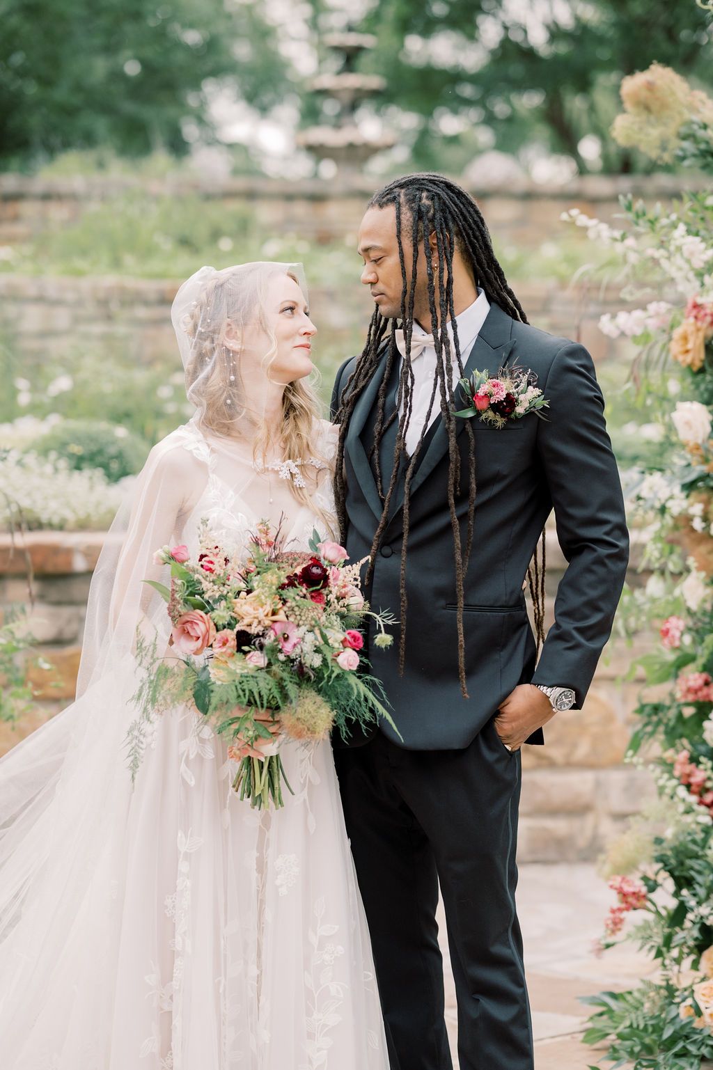 Bride and groom share a romantic moment in a garden, she holds a lush bouquet in a lace gown, he wears a dark suit.