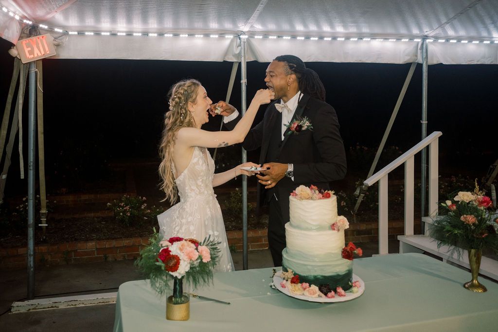 Bride and groom sharing a sweet cake-tasting moment at their wedding reception under a white tent.