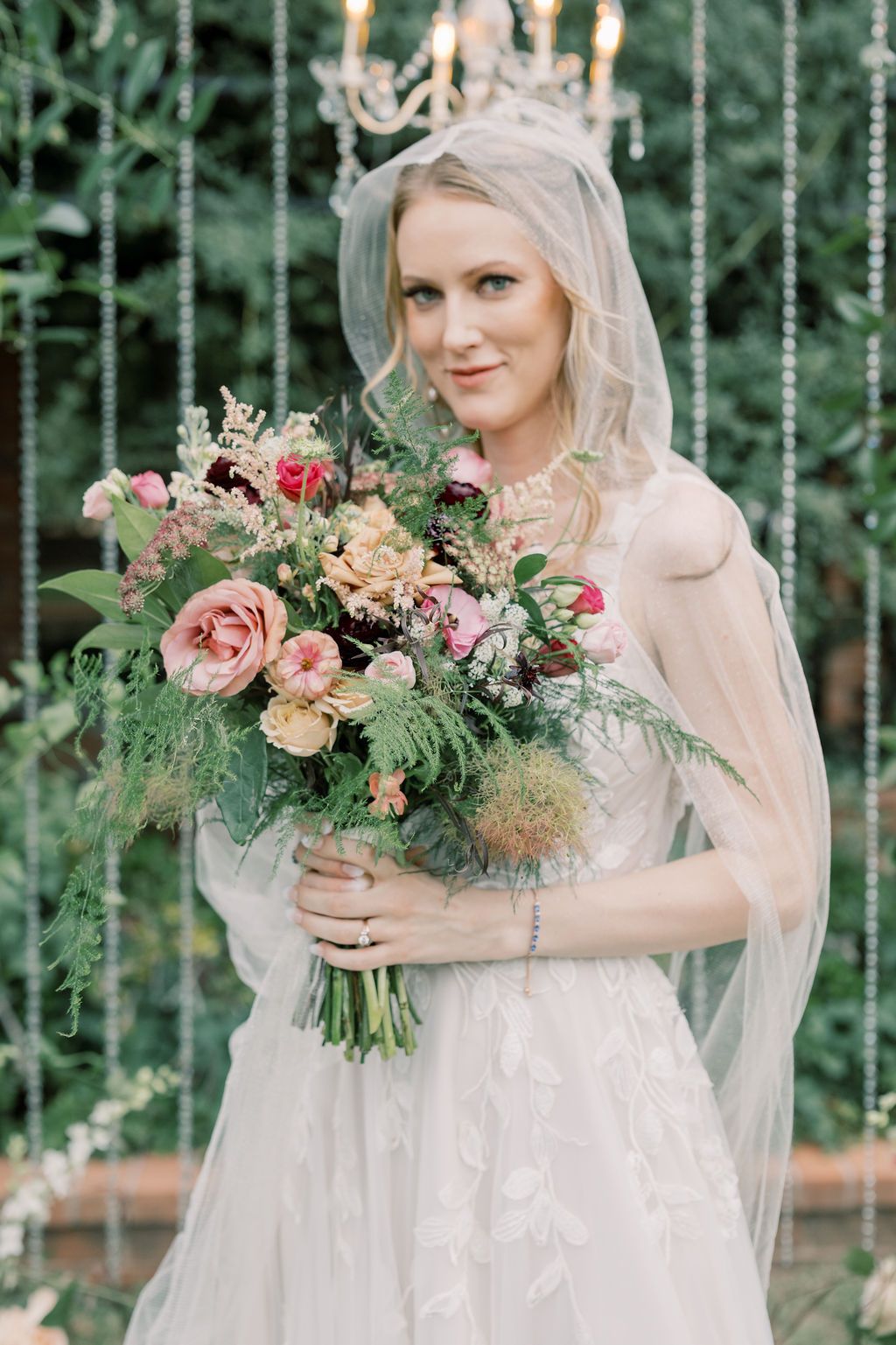 Elegant bride in lace gown and veil holding lush pink and green floral bouquet before crystal chandelier backdrop.