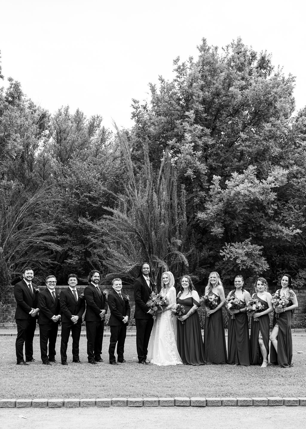 Black and white wedding party photo with bride, groom, groomsmen in suits, and bridesmaids in dark gowns outdoors.