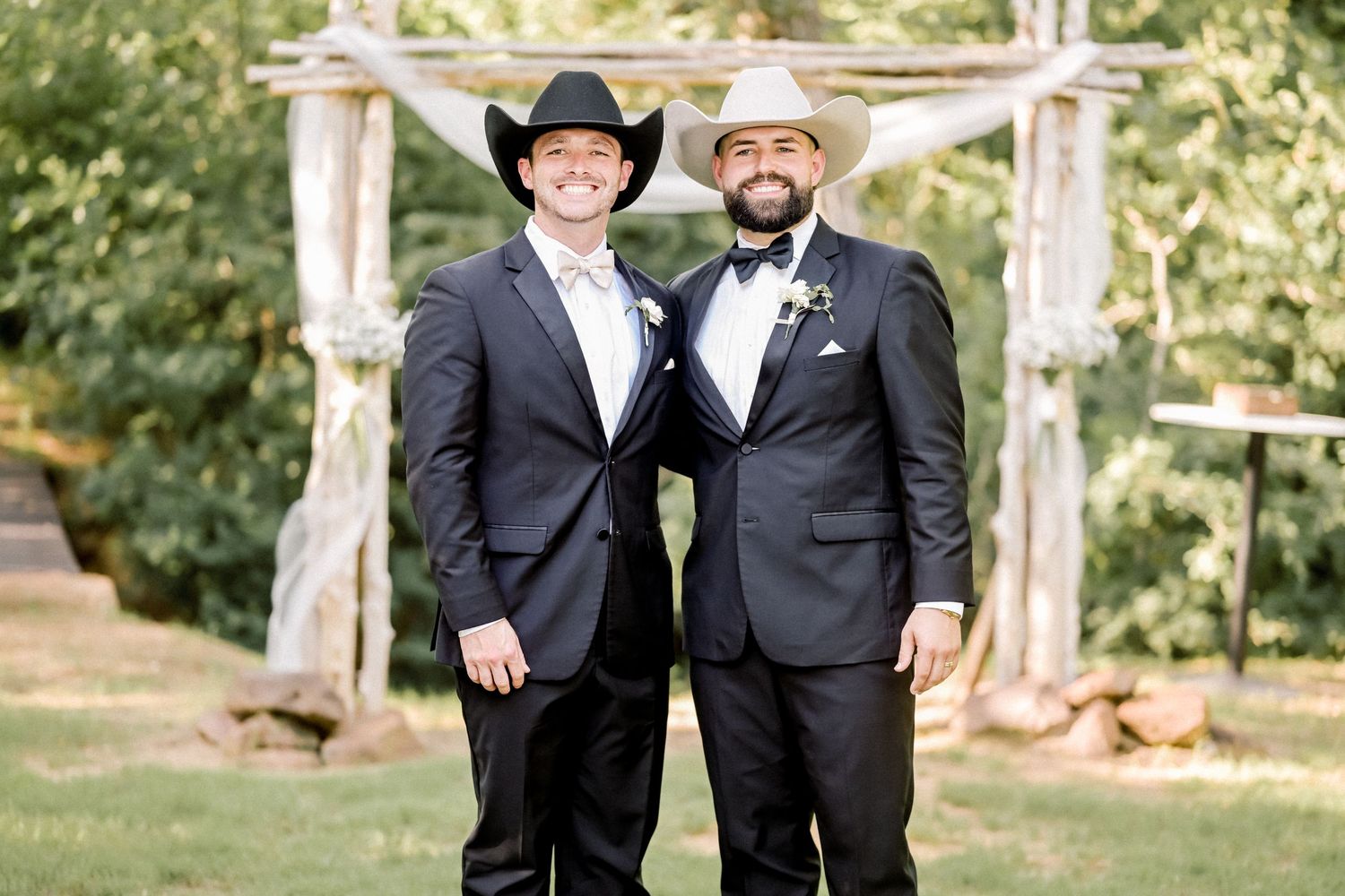 Two groomsmen in black suits and cowboy hats smiling at outdoor rustic wedding ceremony.