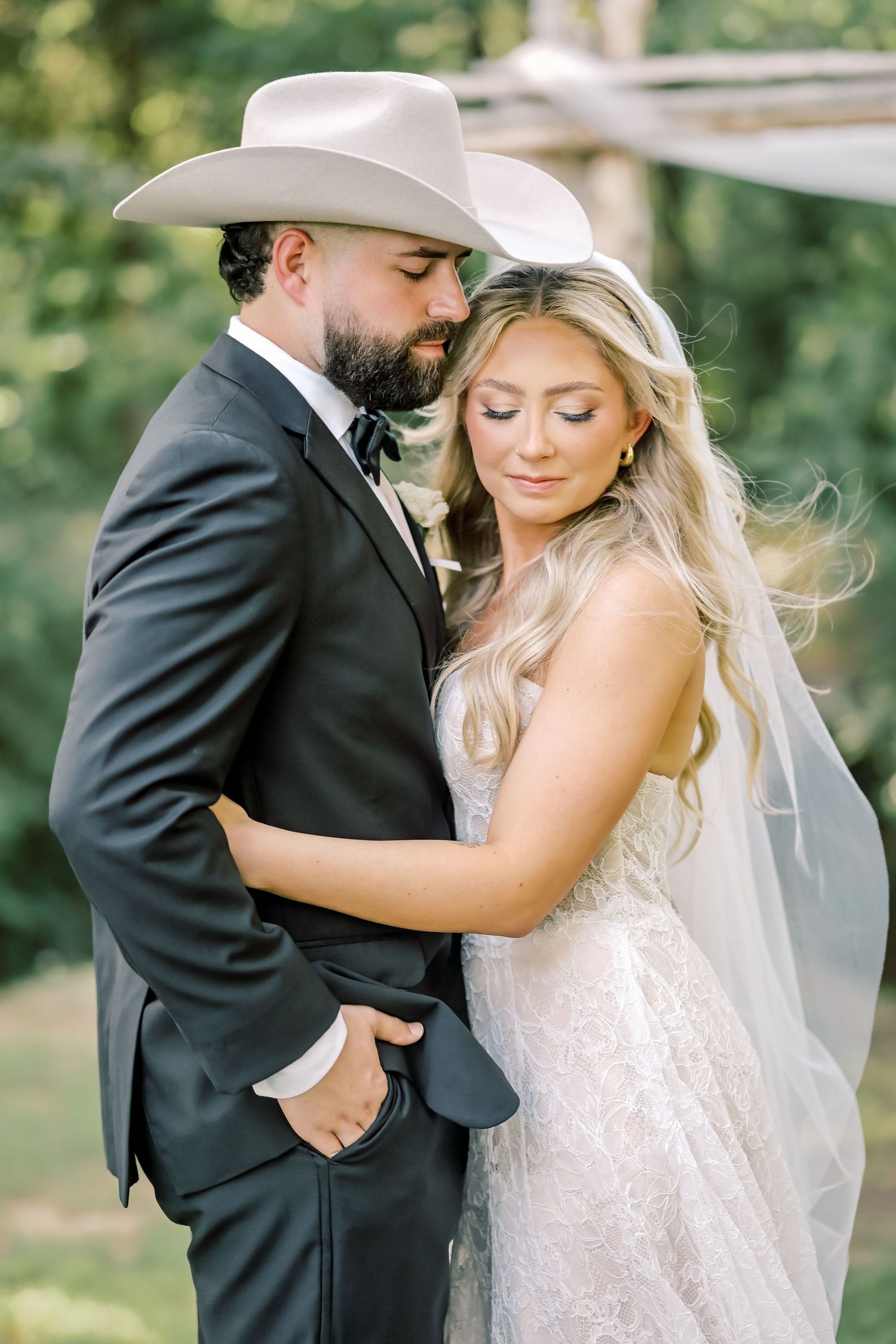 Groom in black suit and white cowboy hat tenderly embraces bride in lace gown outdoors.