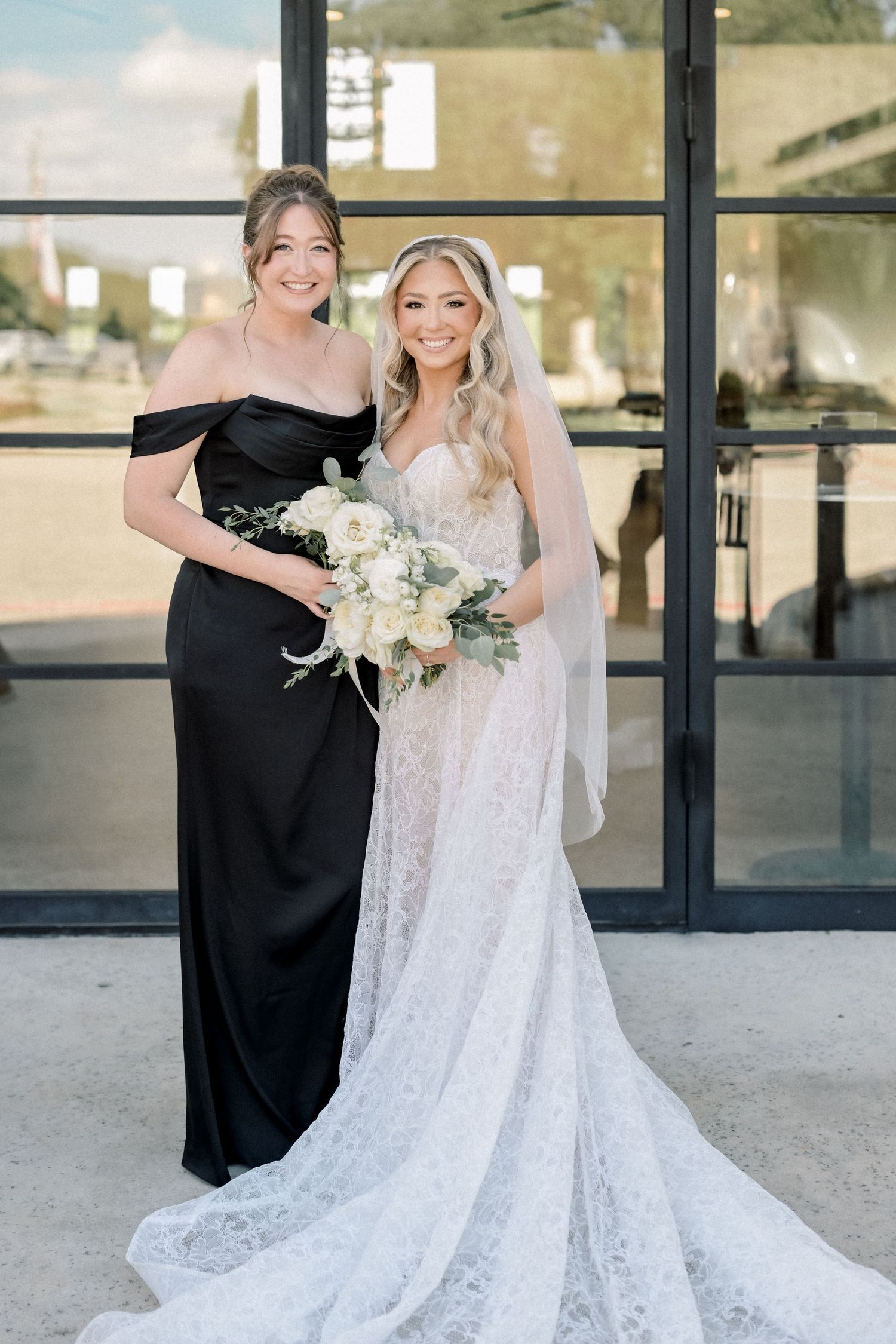 Bride in lace gown holding white bouquet poses with bridesmaid in black off-shoulder dress outdoors.