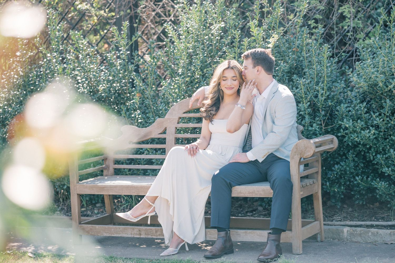 Romantic couple shares tender moments on wooden bench in lush garden setting during golden hour photoshoot.