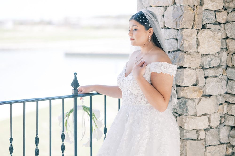 Elegant bride in white lace gown poses against rustic stone wall backdrop.