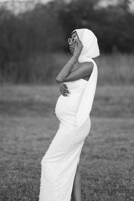 A figure wrapped in white fabric poses gracefully in an outdoor field with trees in the background.