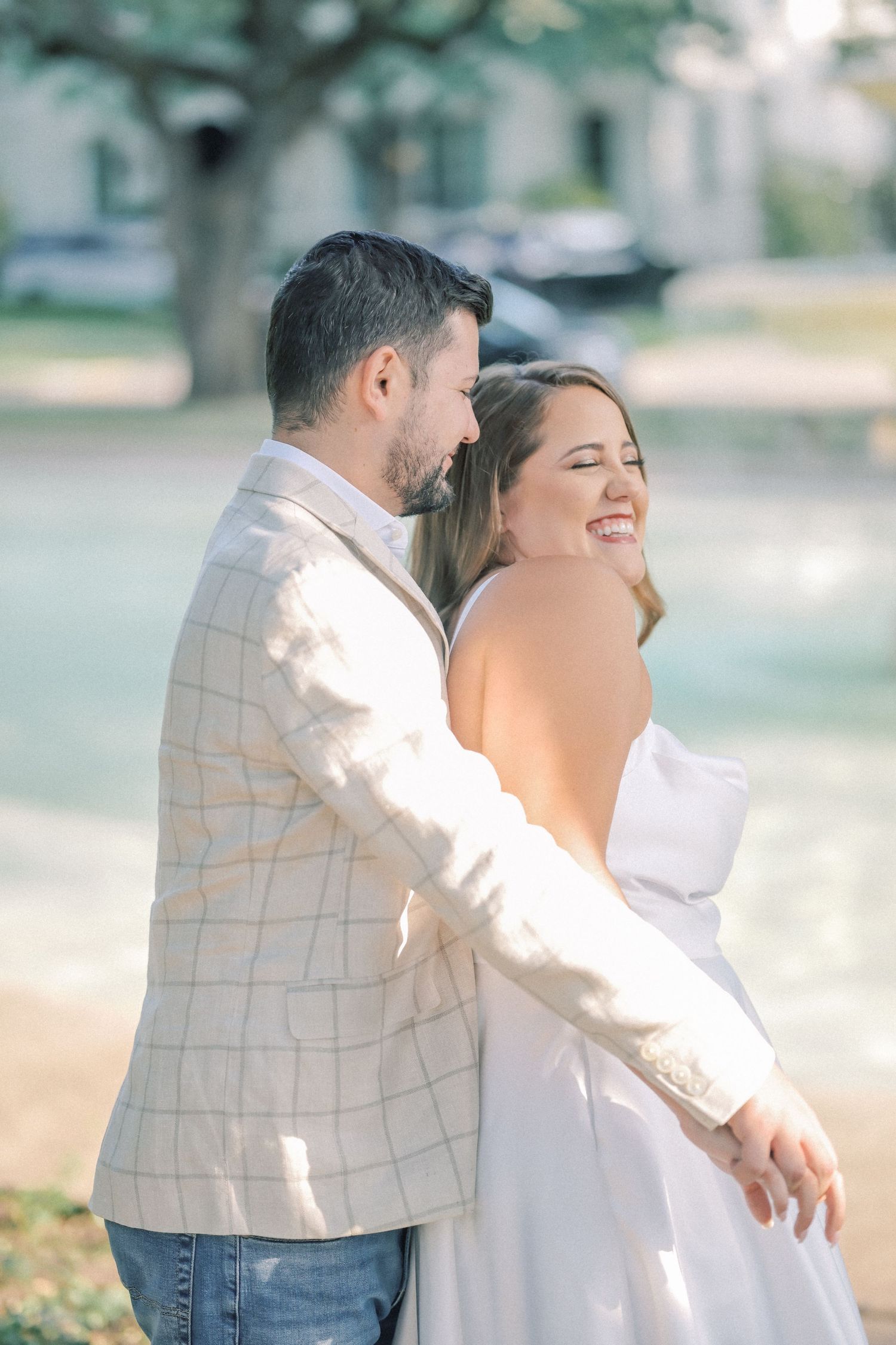 Couple embraces in a sunlit park as the woman laughs with her eyes closed and her fiancé holds her from behind in a plaid blazer.