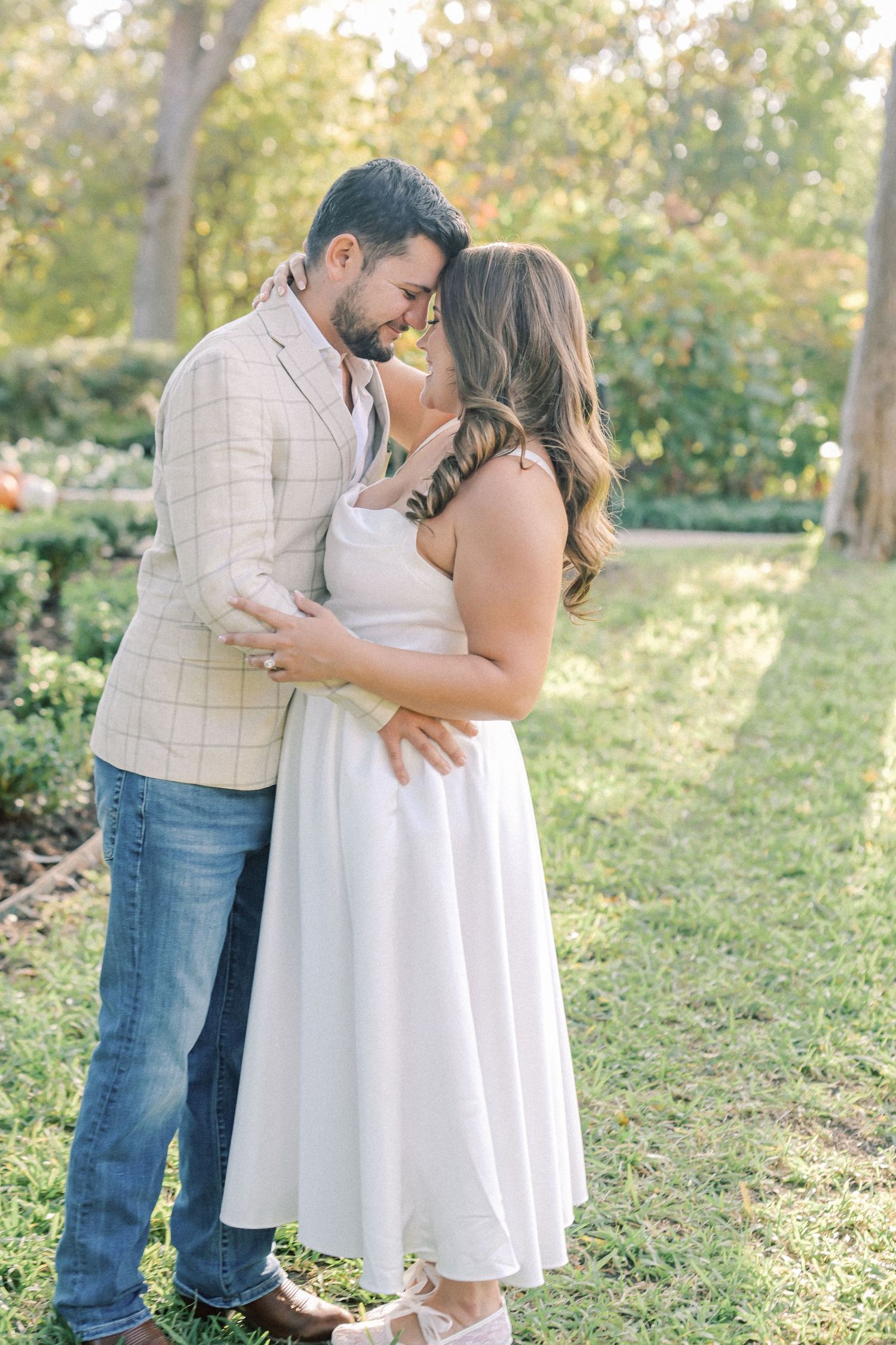 Couple embrace and touch foreheads on a sunlit lawn, she in a flowing white dress and he in a plaid blazer.