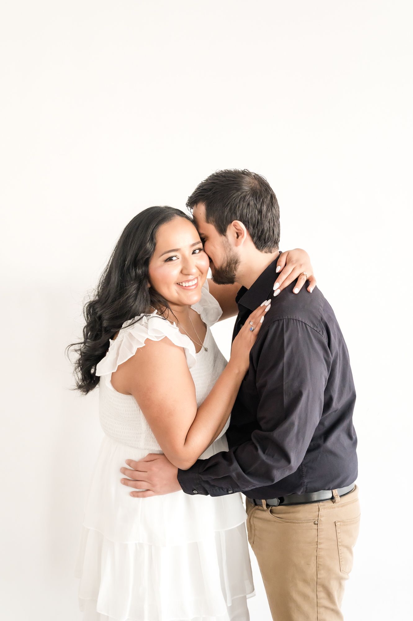 Couple in white and black sharing tender moment against white wall during engagement session.