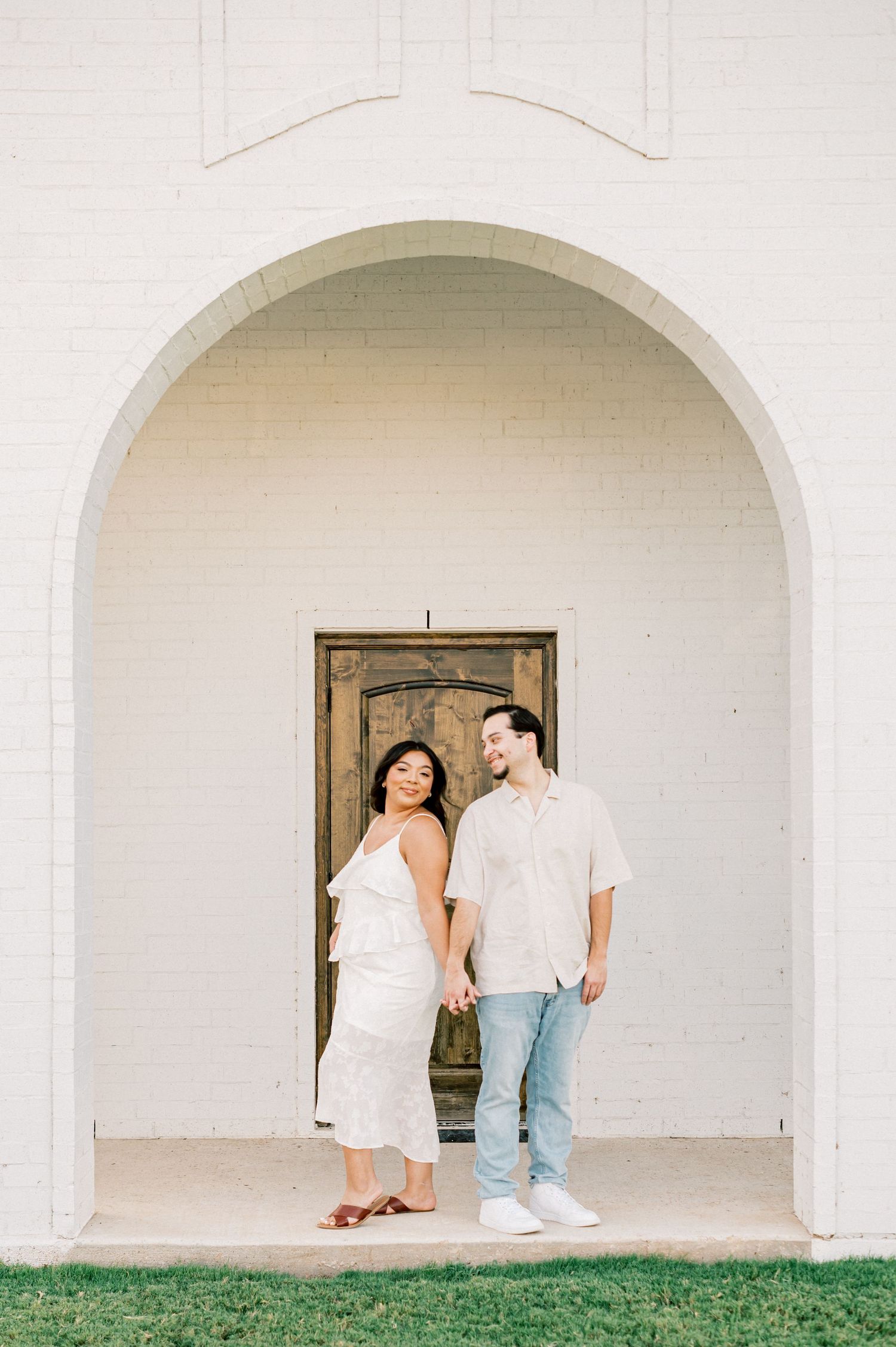 A couple stands together under a white arched doorway wearing light colored outfits on a sunny day.