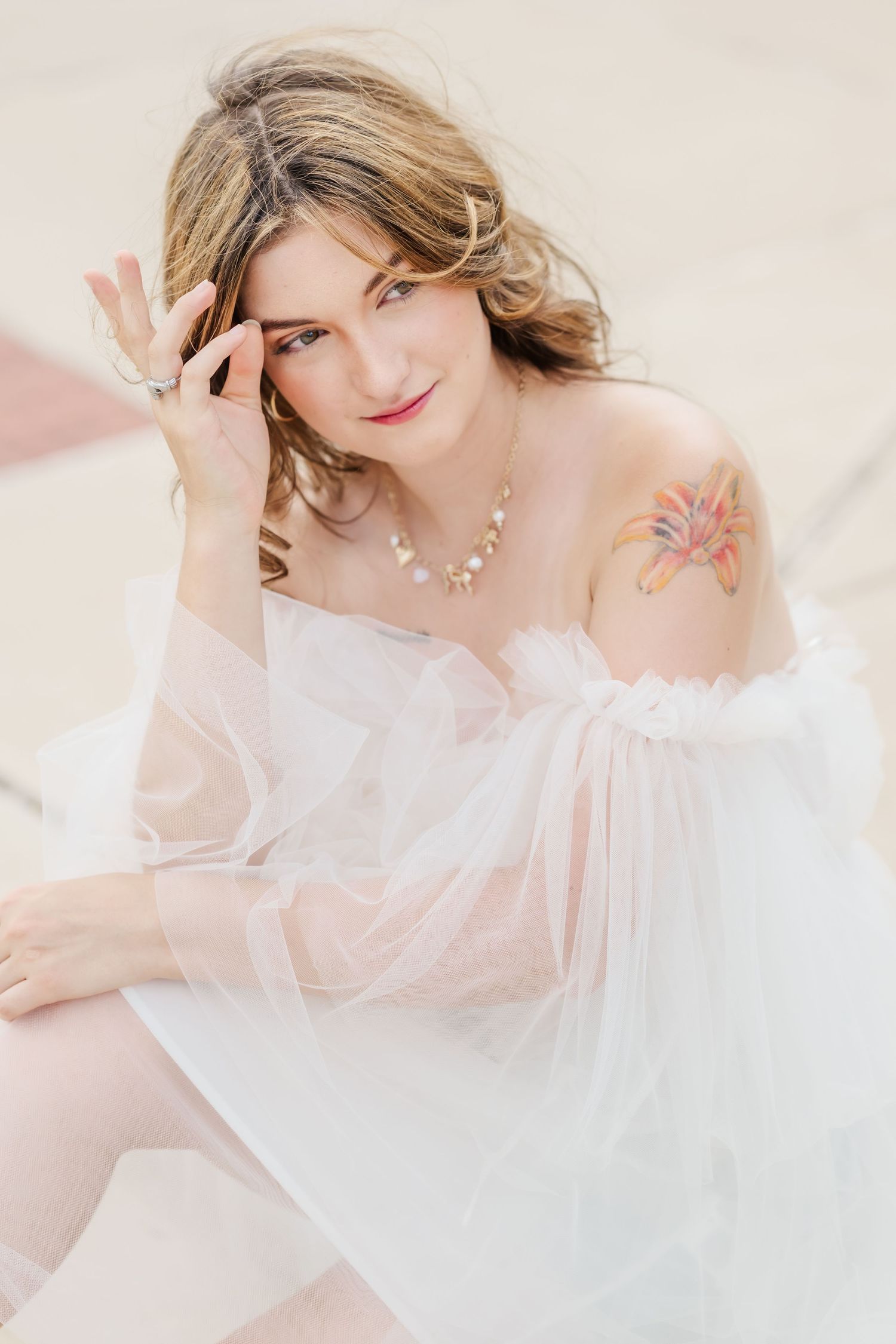 A woman in a sheer white off-shoulder dress with a floral tattoo on her shoulder smiles softly while posing outdoors.
