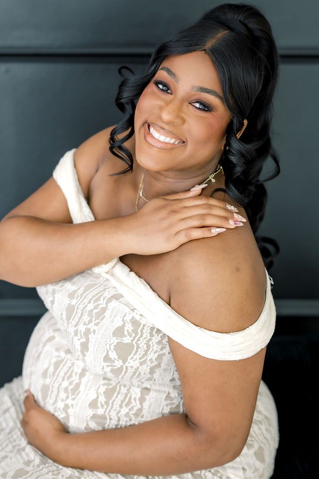 Smiling pregnant woman in white lace off-shoulder dress with elegant updo hairstyle.