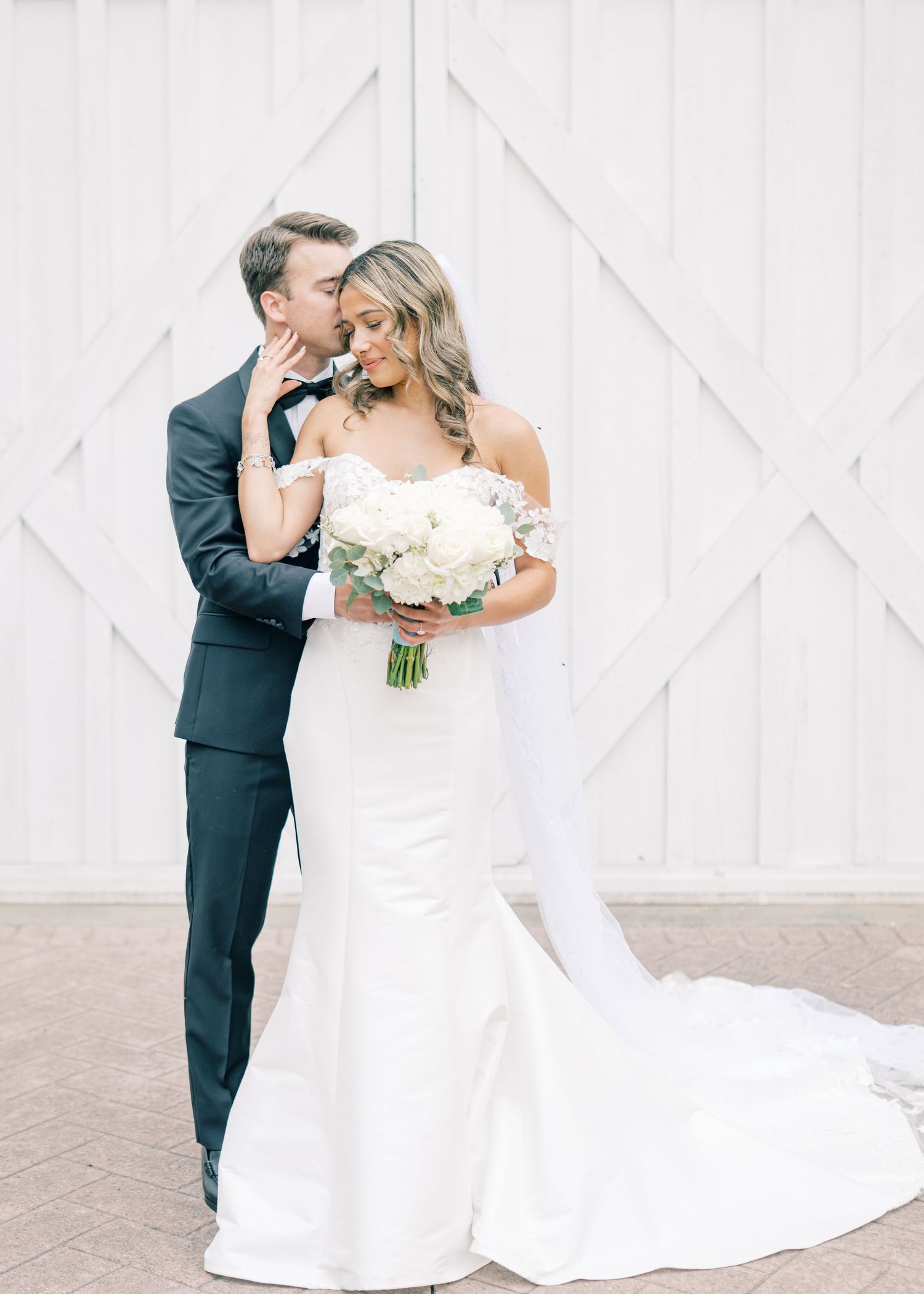 Groom kisses bride holding white floral bouquet in front of barn doors.