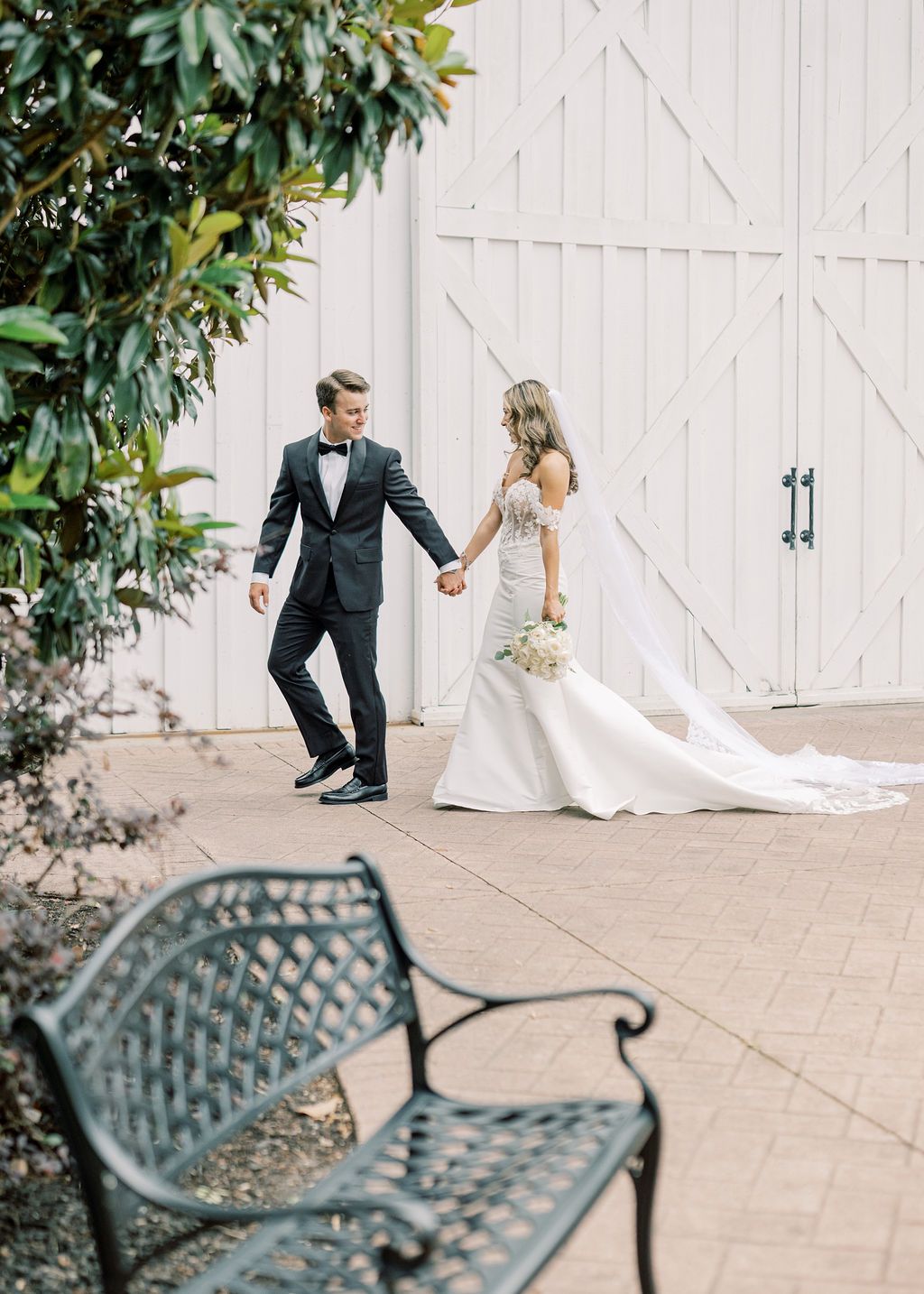 Bride and groom walk hand in hand past white barn, bride in mermaid gown with long train.