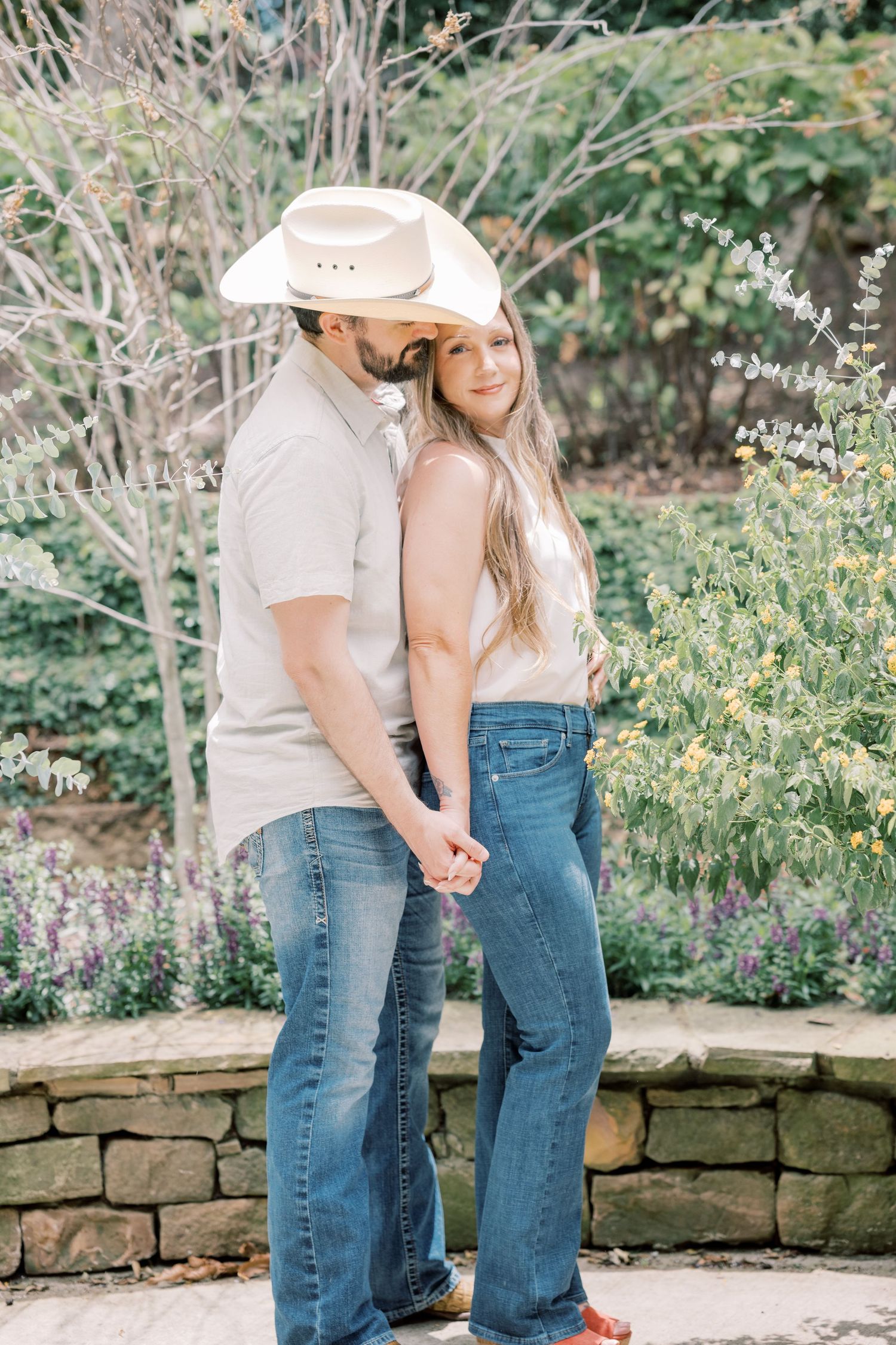 Couple holding hands in a garden, man in white cowboy hat and jeans, woman in flare jeans smiling at camera.