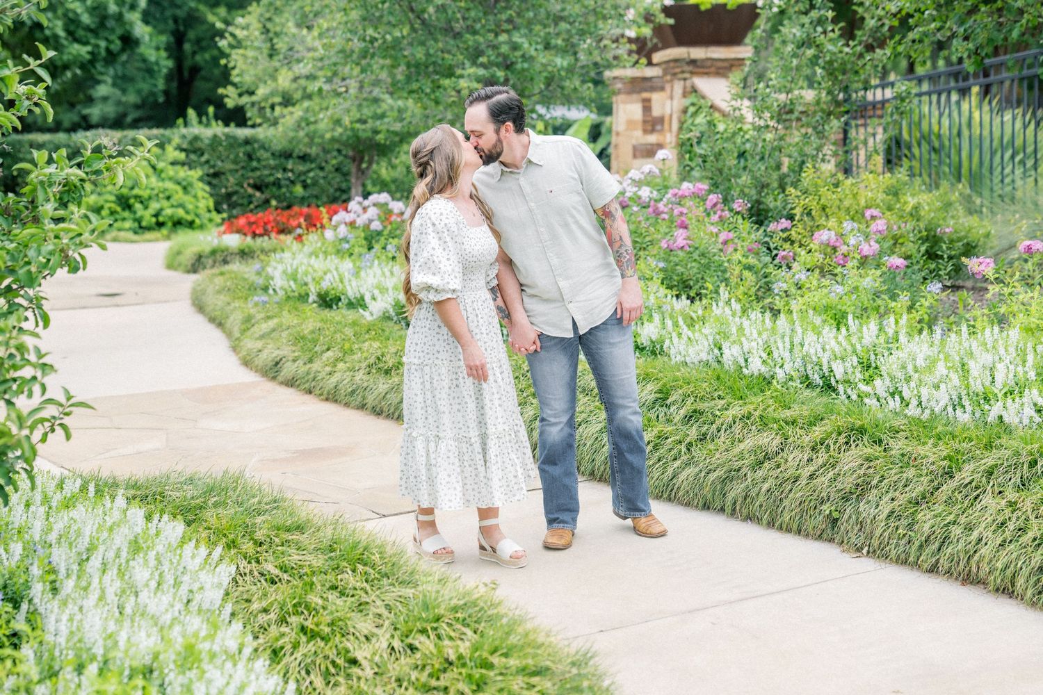 Couple sharing a kiss on a garden path surrounded by lush greenery and colorful flowers in bloom.
