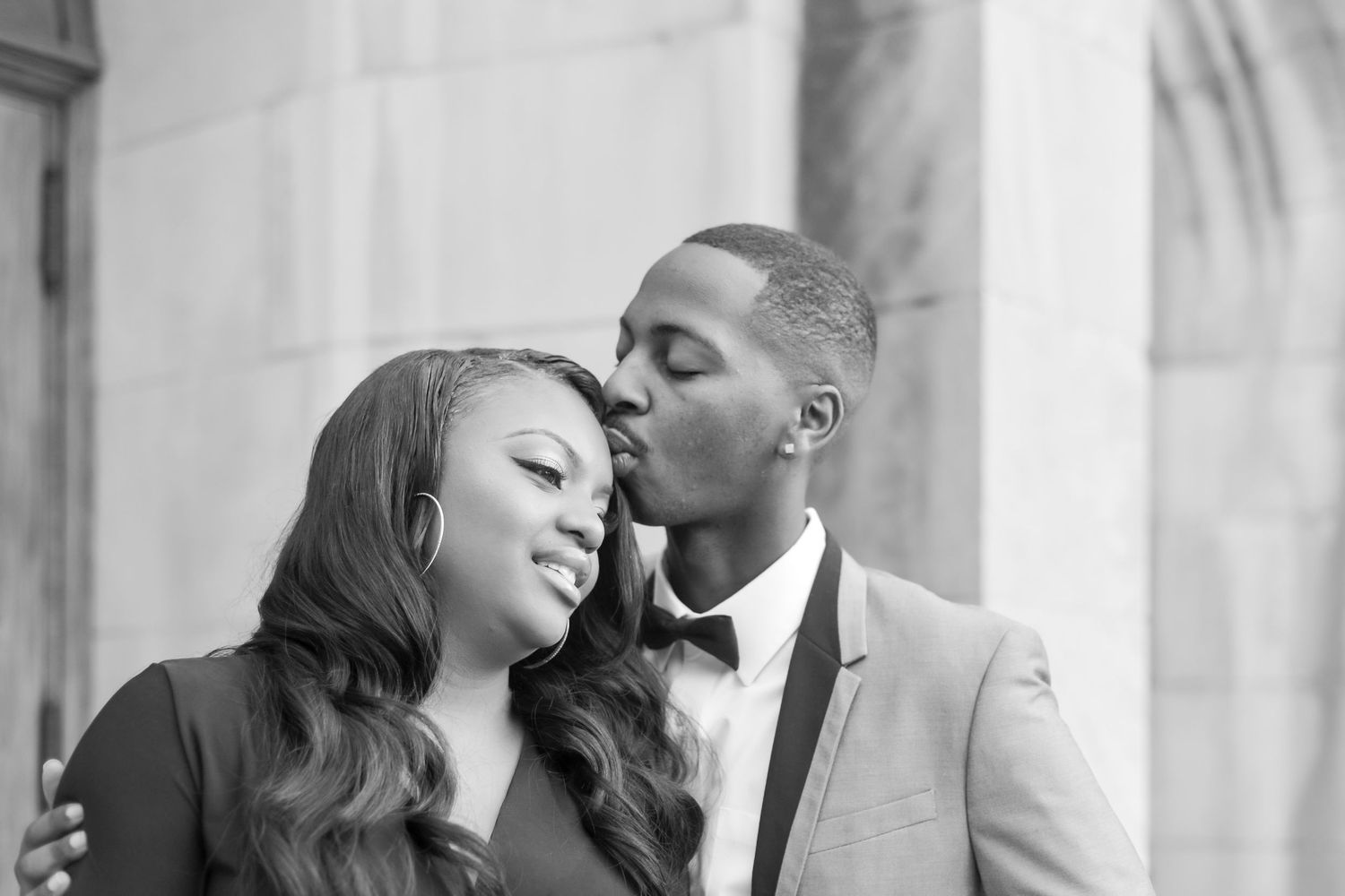 Black and white photo of a couple, man in suit kissing woman on cheek during romantic engagement session.