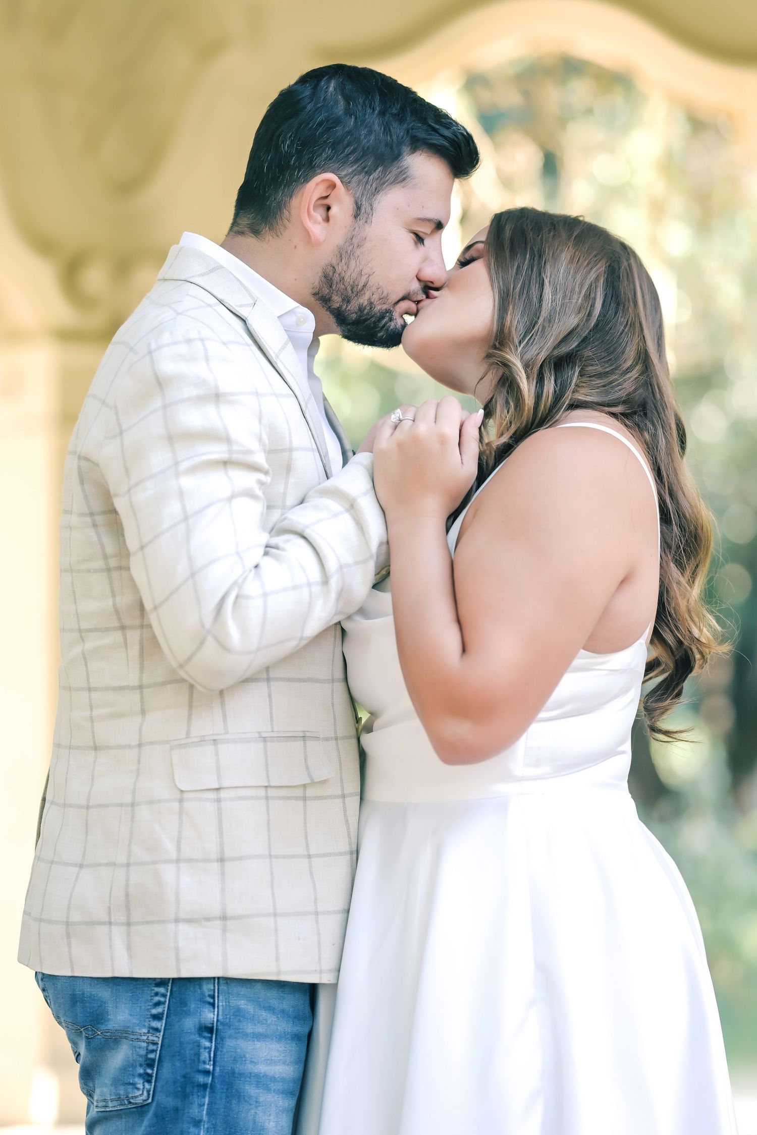 Engaged couple shares a romantic kiss under a stone archway, she in a white dress and he in a plaid blazer and jeans.