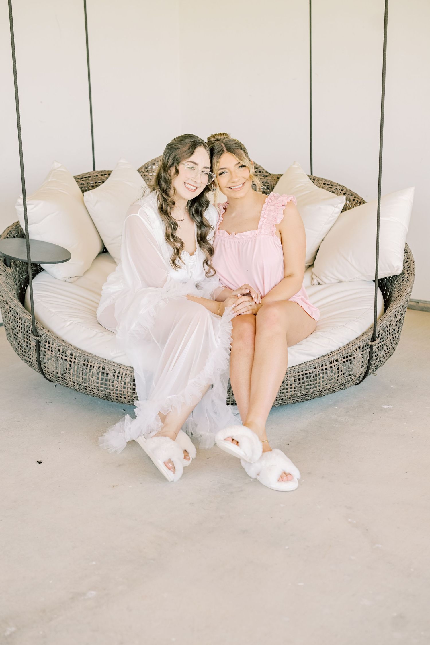 Two smiling women in white and pink outfits sit together on a round wicker swing seat with cream cushions.