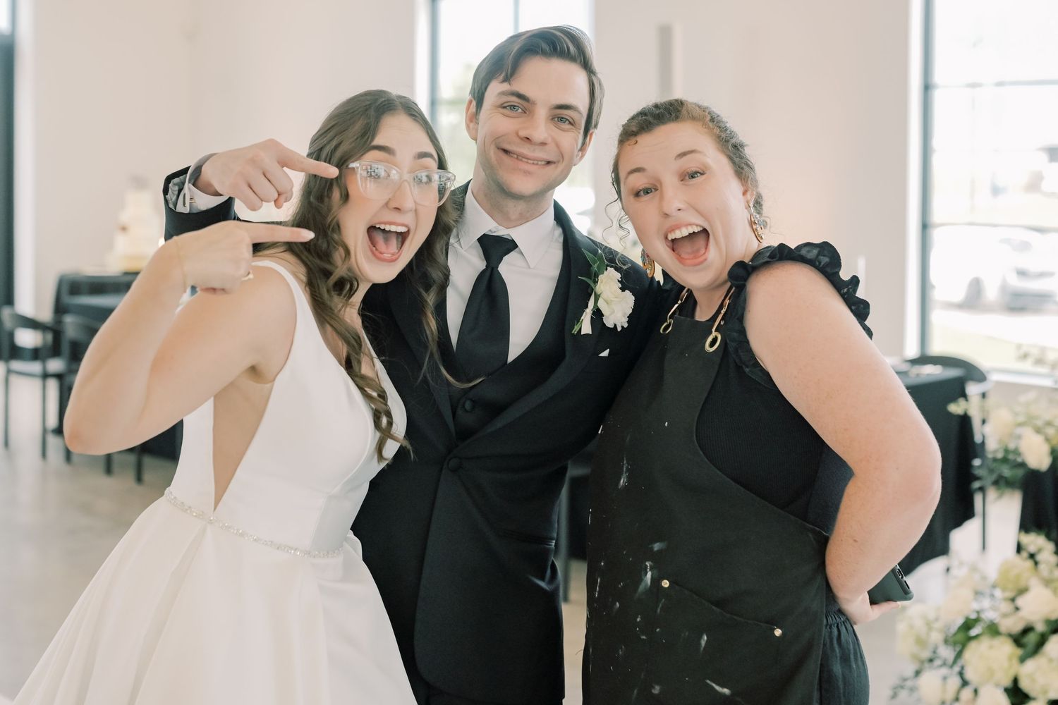 Excited bride, groom in black suit, and guest celebrating joyfully at a wedding reception.