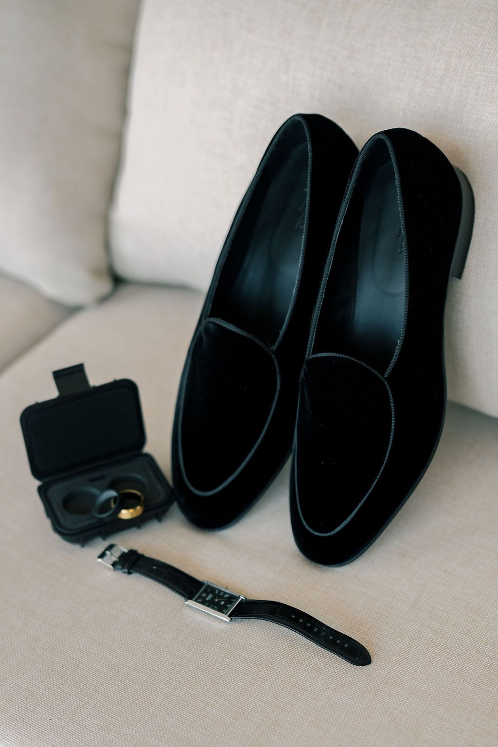 Black velvet loafers with a watch and cufflinks displayed on a beige chair for a wedding day.