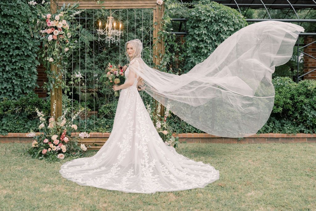 Bride in lace wedding gown with flowing veil outdoors near floral arch and greenery backdrop.