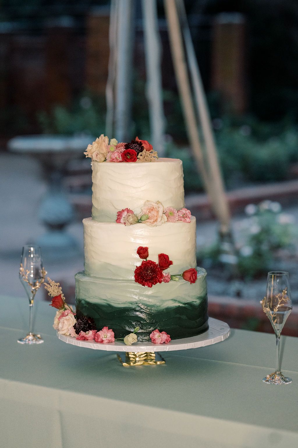 Three-tier wedding cake with green ombre frosting and fresh floral decorations, displayed on elegant cake stand.