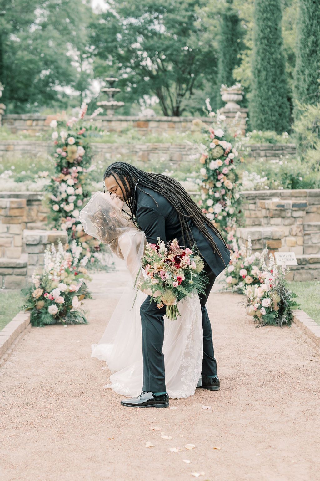 Bride and groom share a romantic dip kiss at an outdoor garden wedding ceremony with floral arrangements.